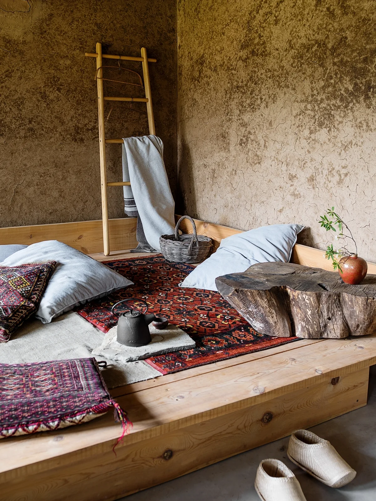 A low wooden platform bed features patterned rugs, cushions, a live-edge side table, and a leaning ladder against textured plaster walls.