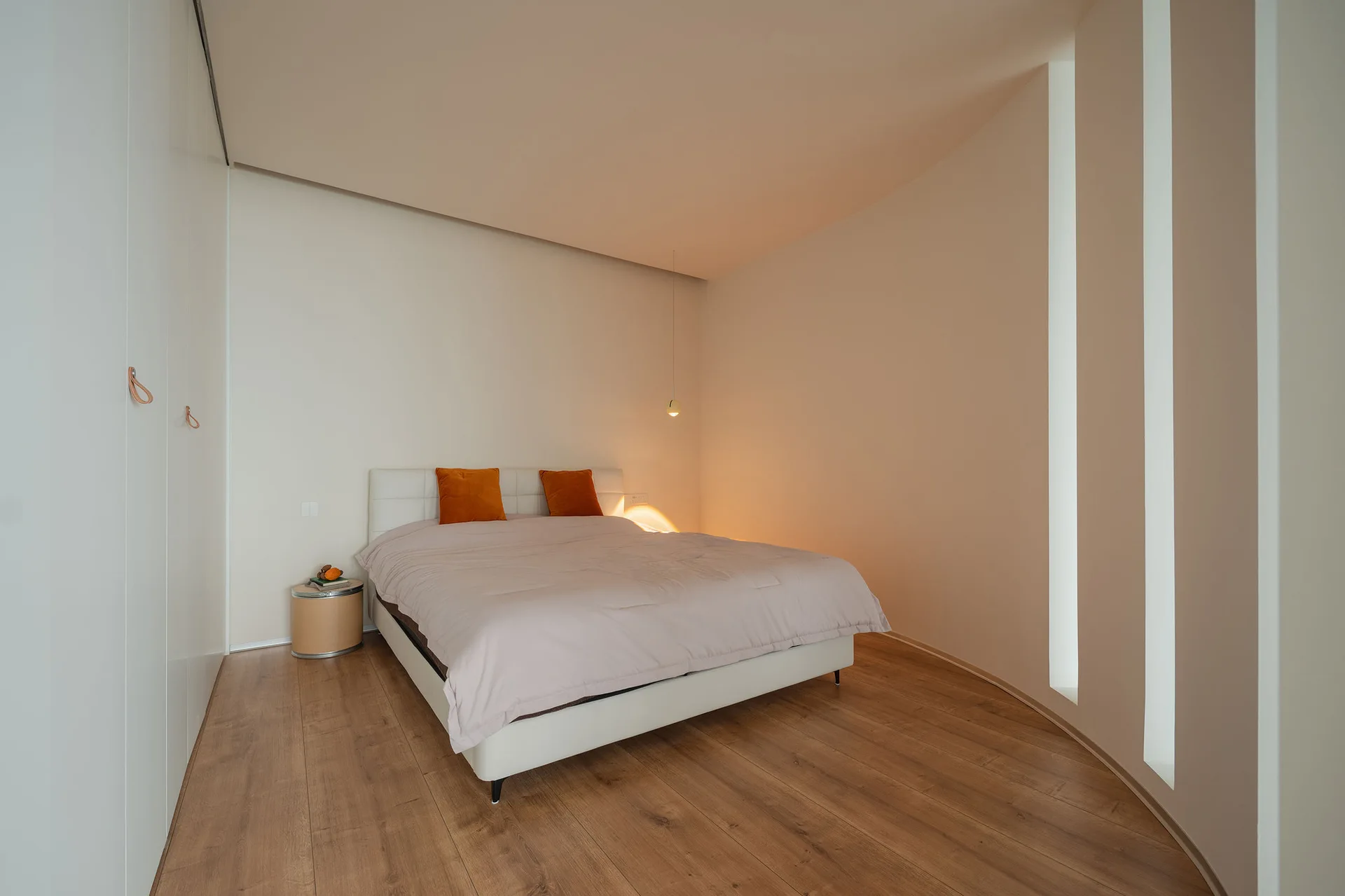 A minimalist bedroom features a bed with orange pillows, wood flooring, built-in white wardrobes, and three vertical slit windows on a curved wall.