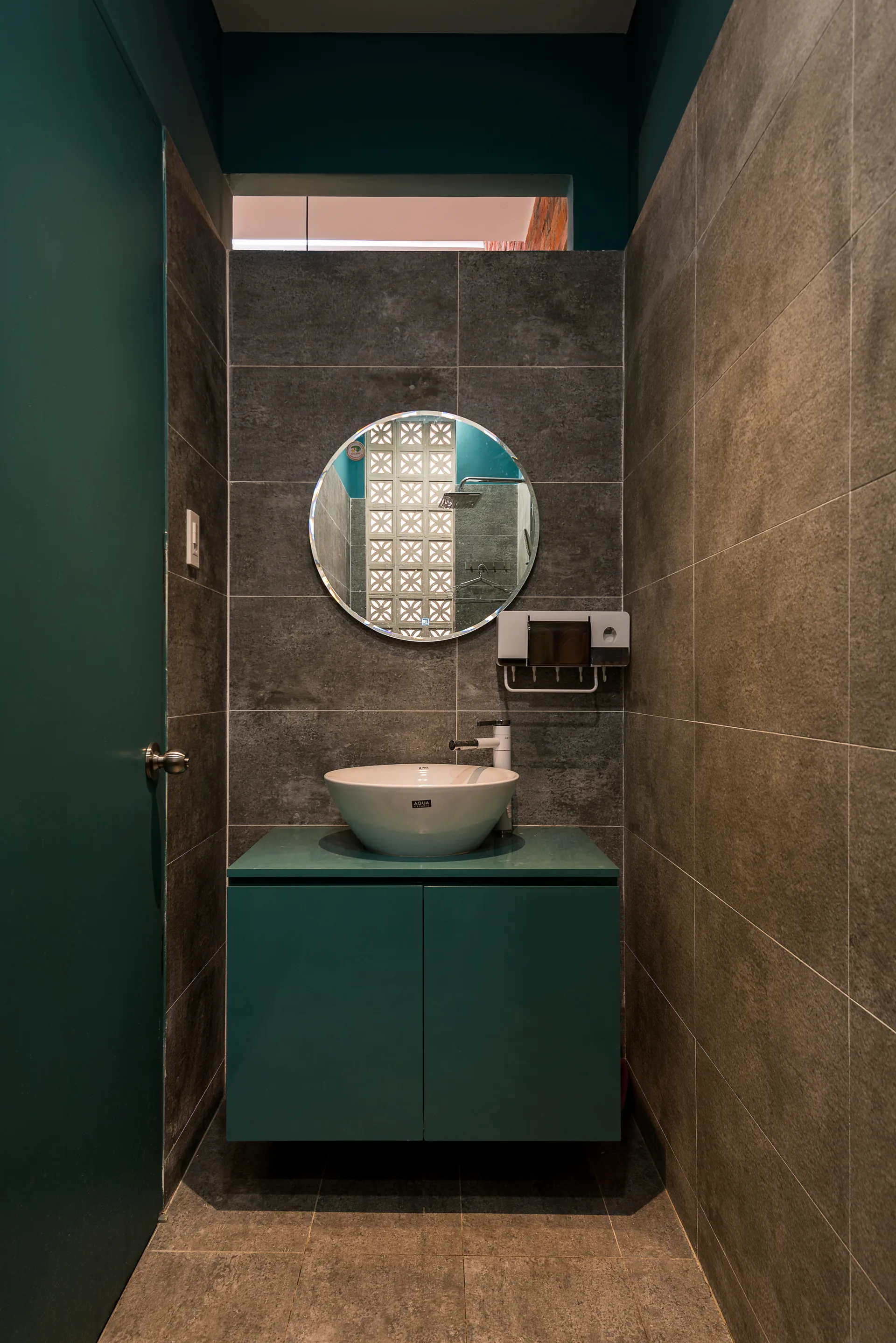 A minimalist bathroom features a floating teal cabinet, a white vessel sink, a circular mirror, and textured grey wall tiles.