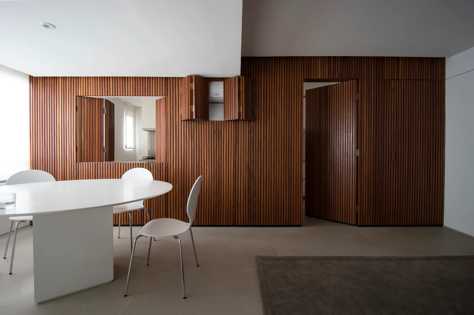 A white oval dining table and matching chairs sit on a light tile floor next to a wall covered in vertical wooden slats with built-in doors.