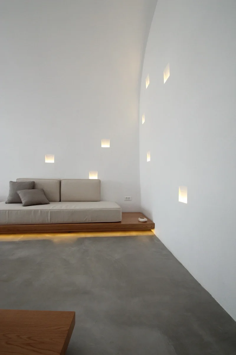 A low-profile beige platform sofa rests on a wooden base next to a white plaster wall featuring glowing square recessed light niches and a gray concrete floor.