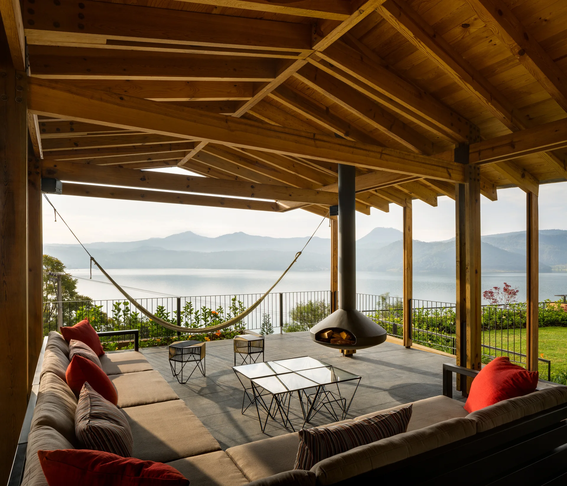 A covered lakeside patio features a large brown sectional sofa, a black suspended fireplace, and a natural wood beam ceiling looking out at a lake.