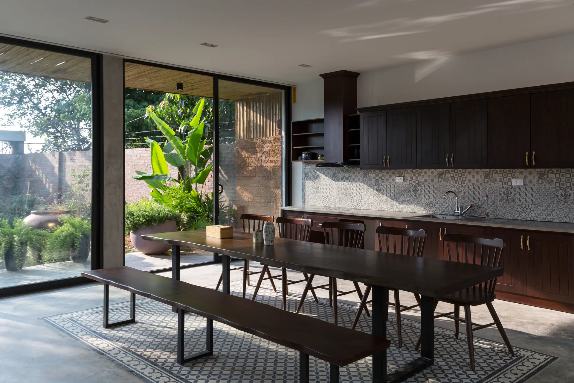 A kitchen featuring dark wood cabinetry, a patterned backsplash, and a long wooden dining table on a concrete floor with a tile inlay near sliding glass doors.