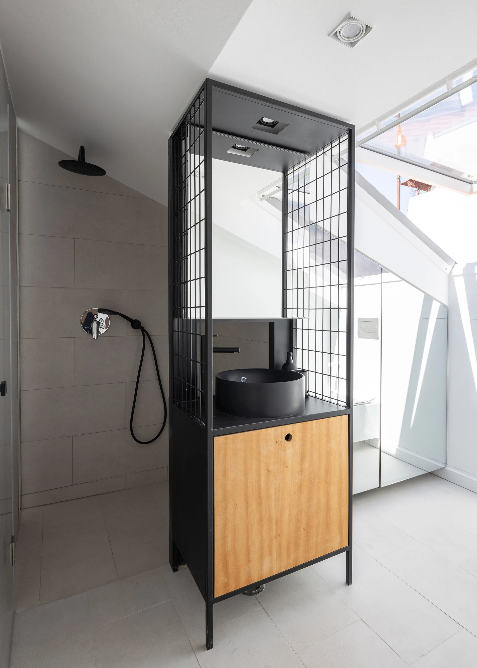 A modern attic bathroom featuring a black metal wireframe vanity with a round vessel sink under a sloped ceiling skylight.