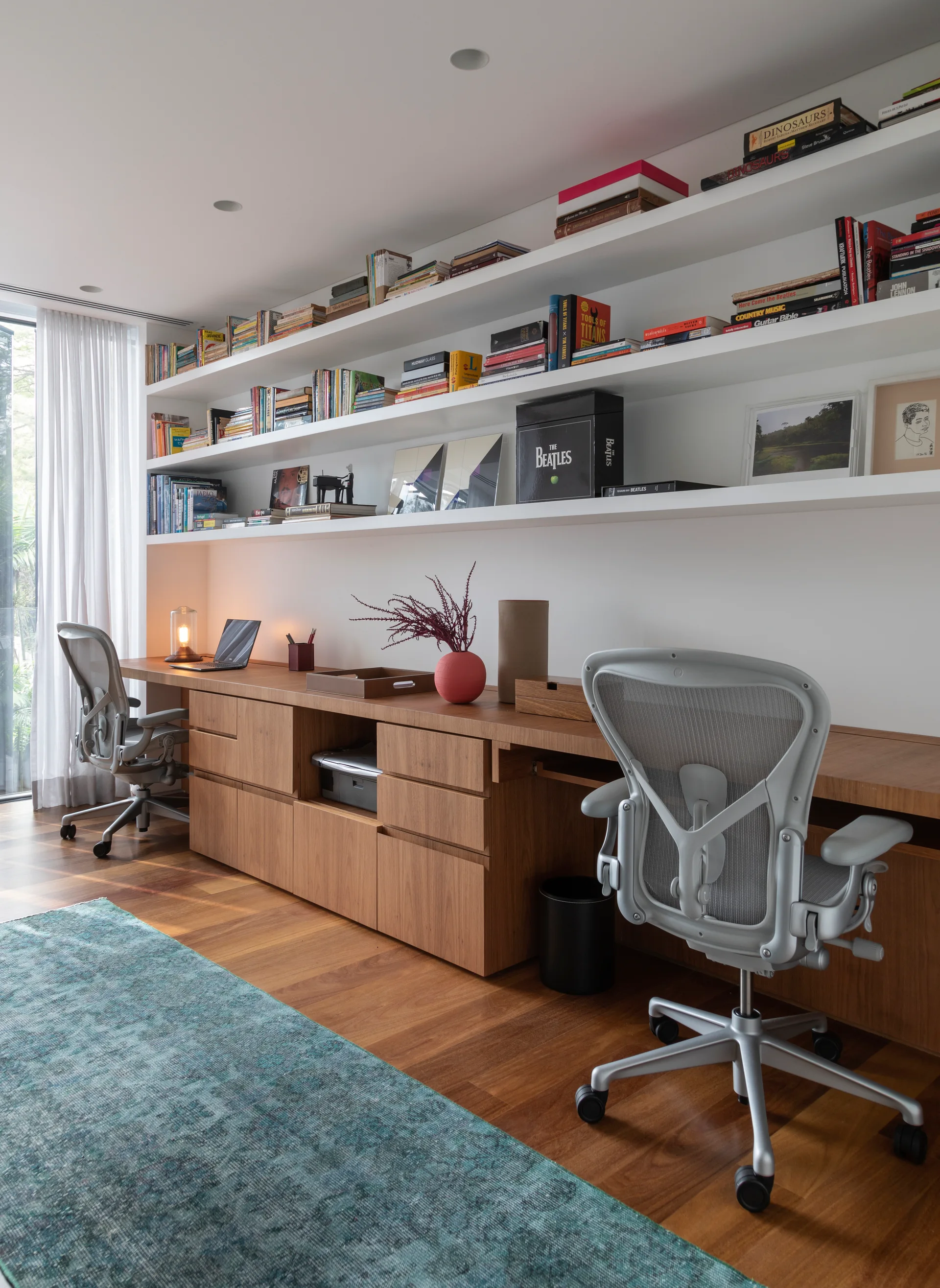A modern home office featuring a long built-in wooden desk with two ergonomic mesh chairs and white wall-mounted shelves filled with books.