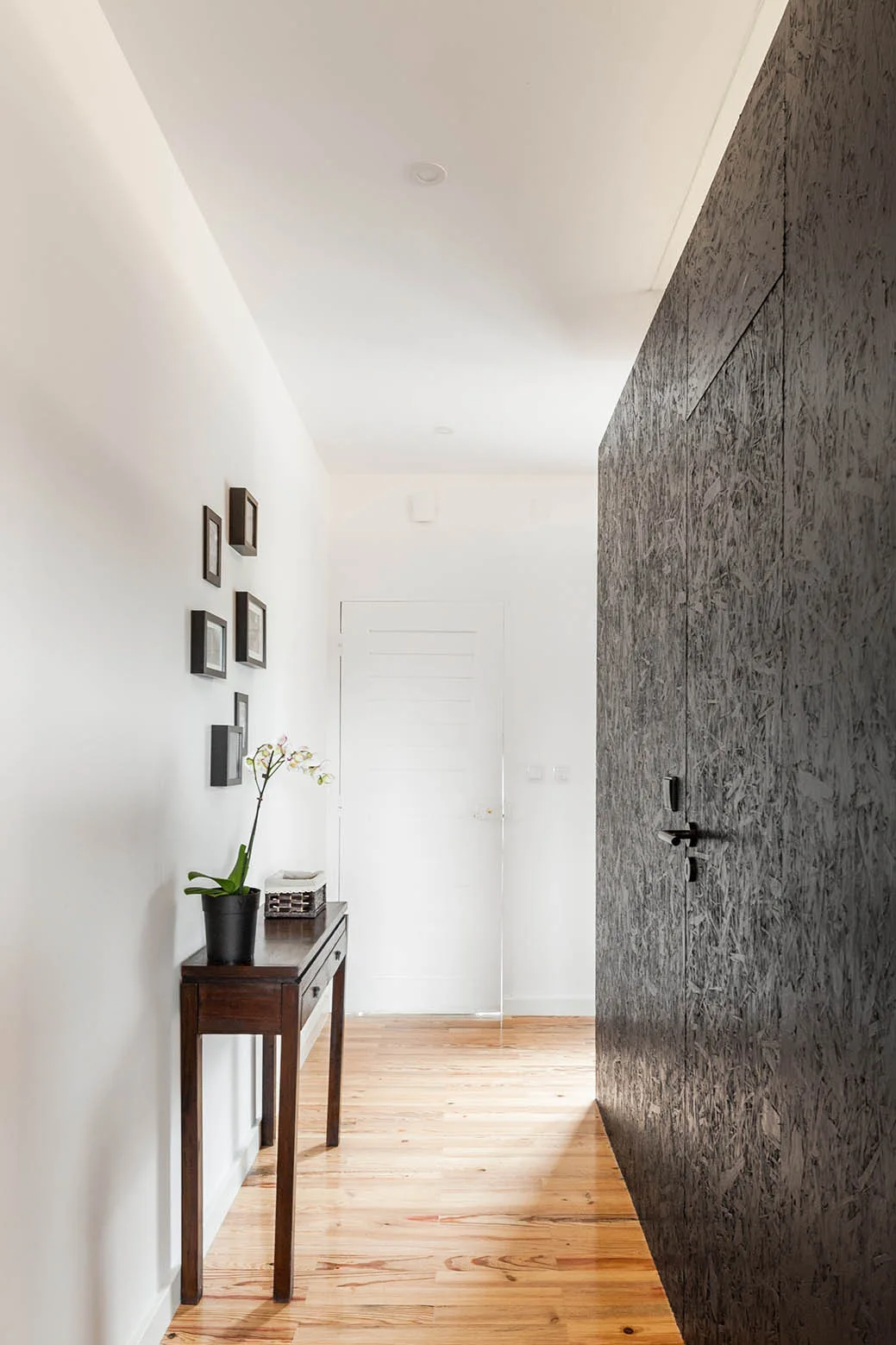 A narrow hallway features a dark stained OSB panel wall on one side, a slim dark console table with a plant on the other, and natural wood flooring.