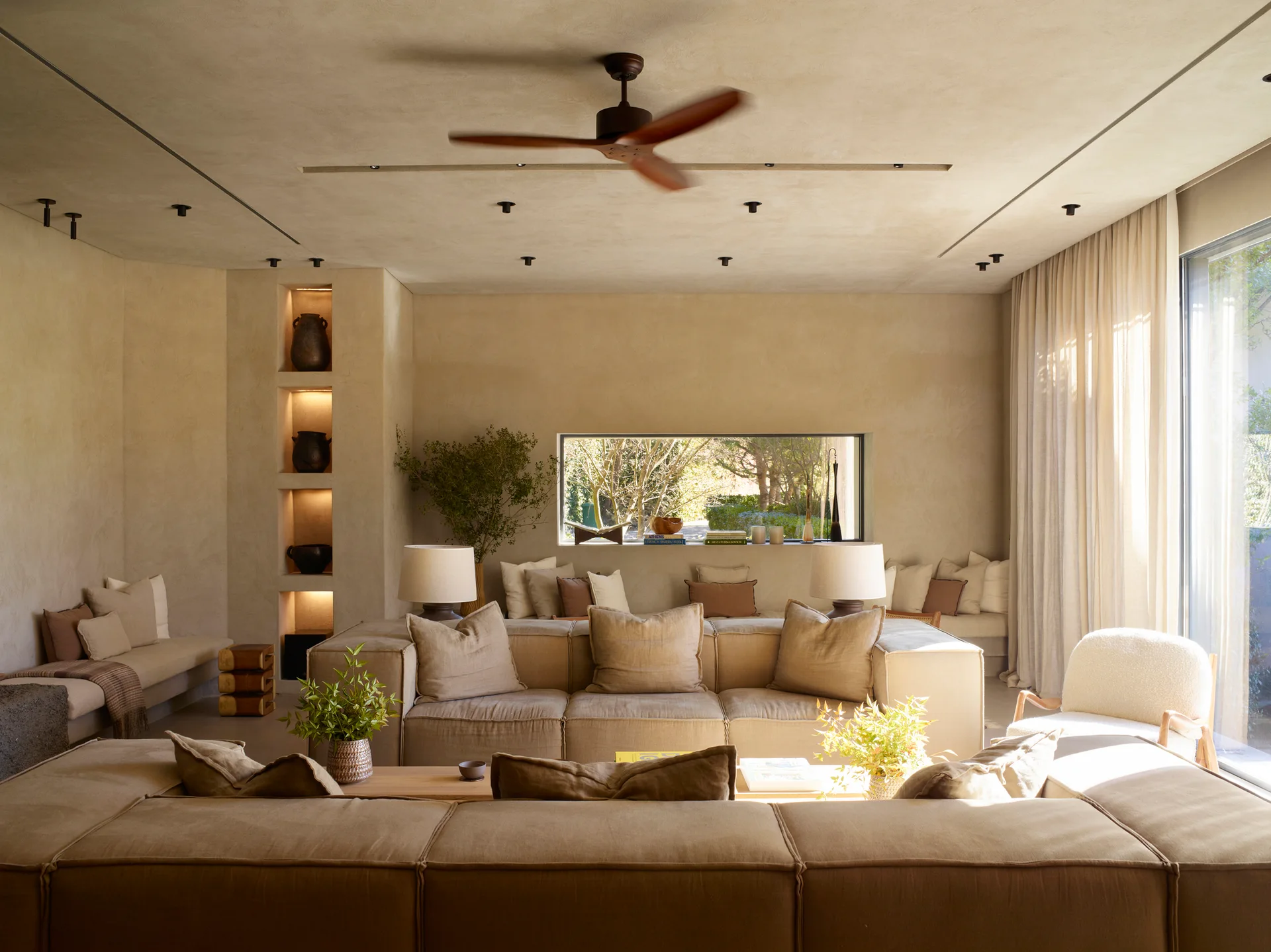 A beige plastered living room with a large modular sofa, vertical lit wall niches containing vases, and a horizontal window behind perimeter benches.
