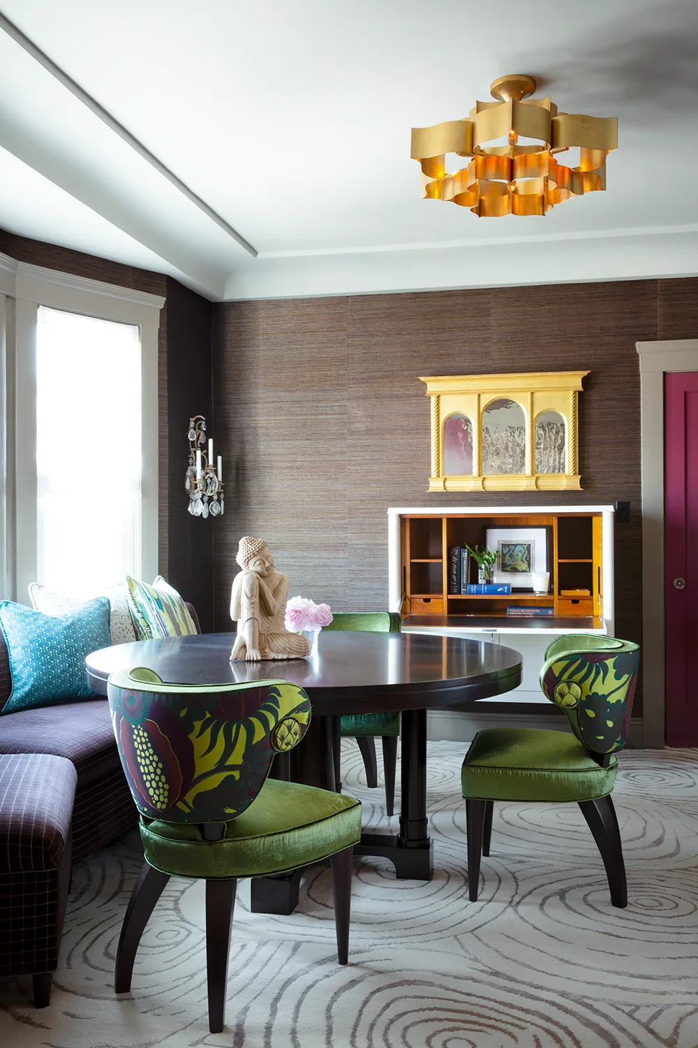 An eclectic dining space features a dark wood table, green velvet chairs with patterned backs, dark brown textured wallpaper, and a gold ceiling fixture.