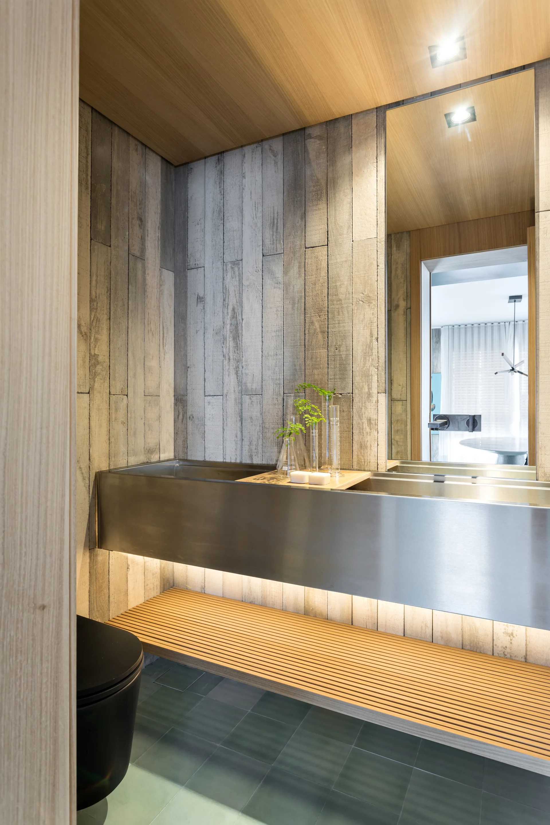 A modern powder room features a floating stainless steel trough sink, a lower wooden slatted shelf, a black toilet, and gray wood-grain paneled walls.