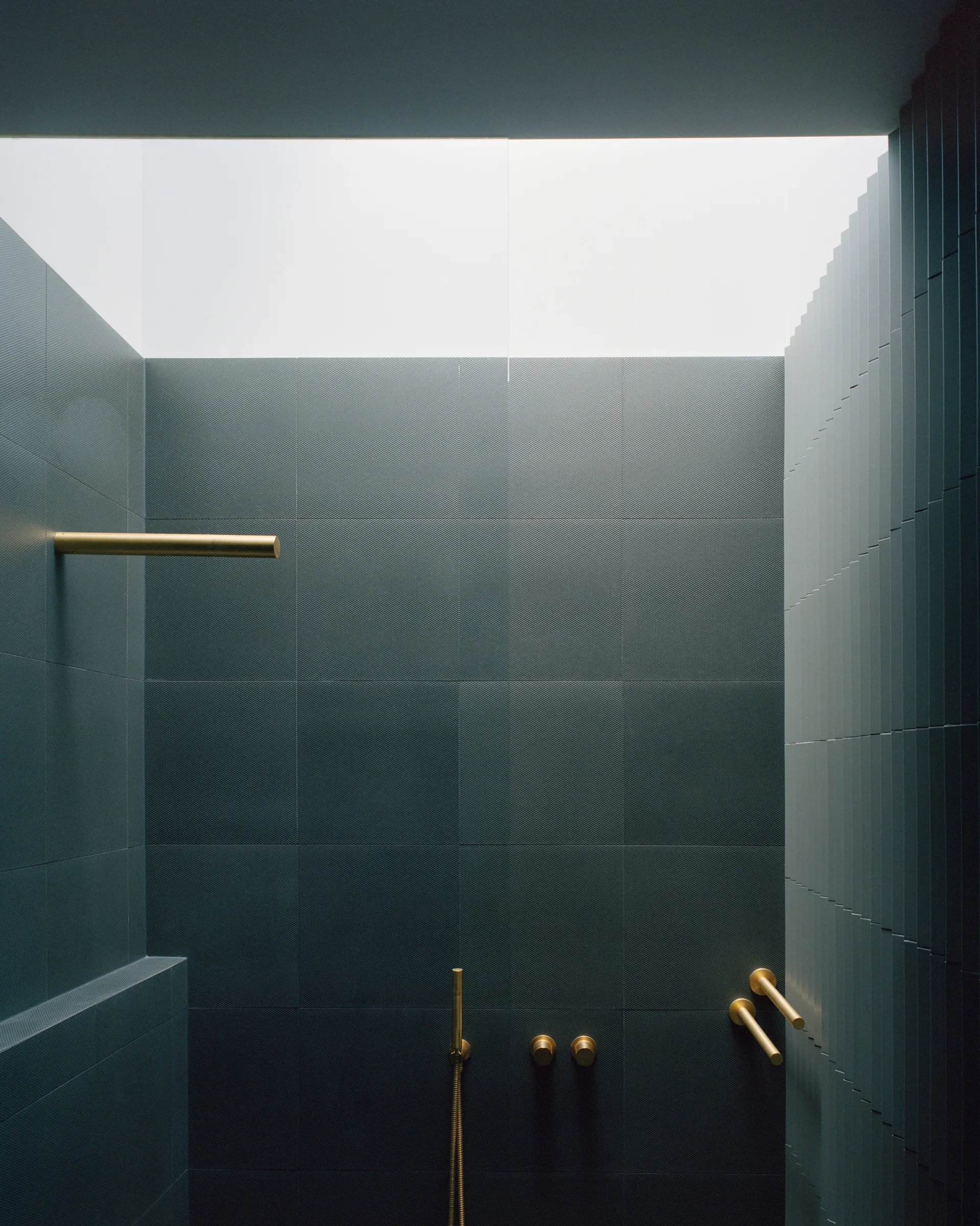 A minimalist shower stall featuring dark charcoal grid tiles, brushed brass fixtures, and natural light streaming down from an overhead skylight.