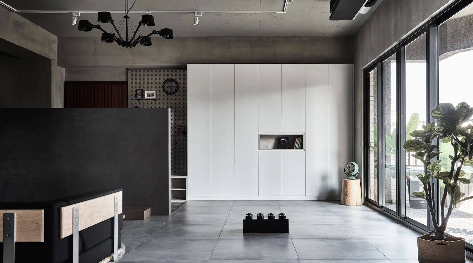 A minimalist living room features large white built-in cabinets against concrete walls and floors, with a black room divider and sliding glass doors.