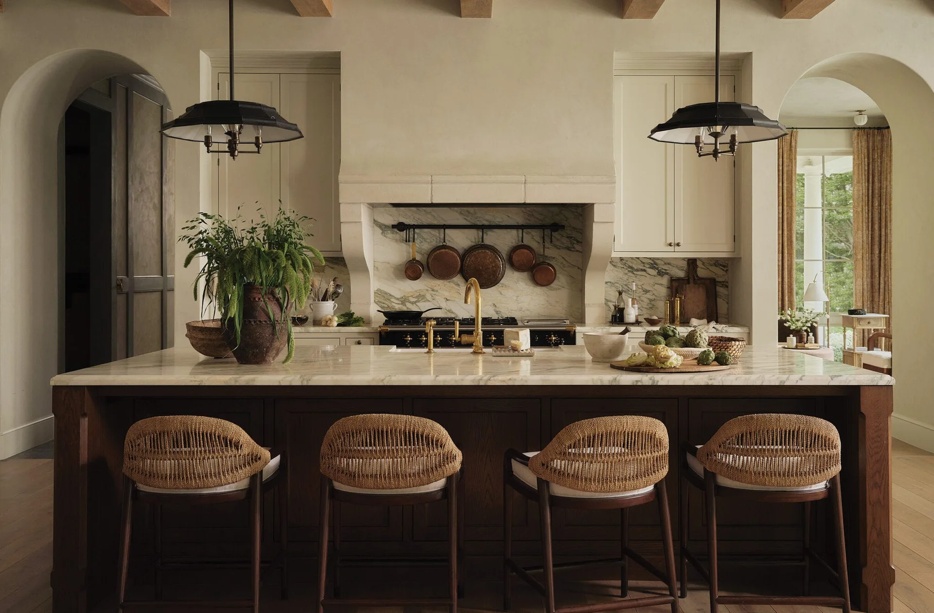A rustic kitchen features a marble-topped dark wood island with four woven bar stools, cream plaster walls, and hanging copper pans.