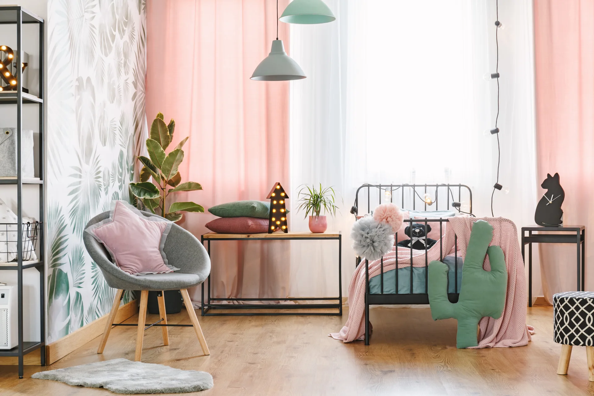 A children's bedroom features a black metal bed, pink curtains, green botanical wallpaper, a grey armchair, and playful decorative pillows.