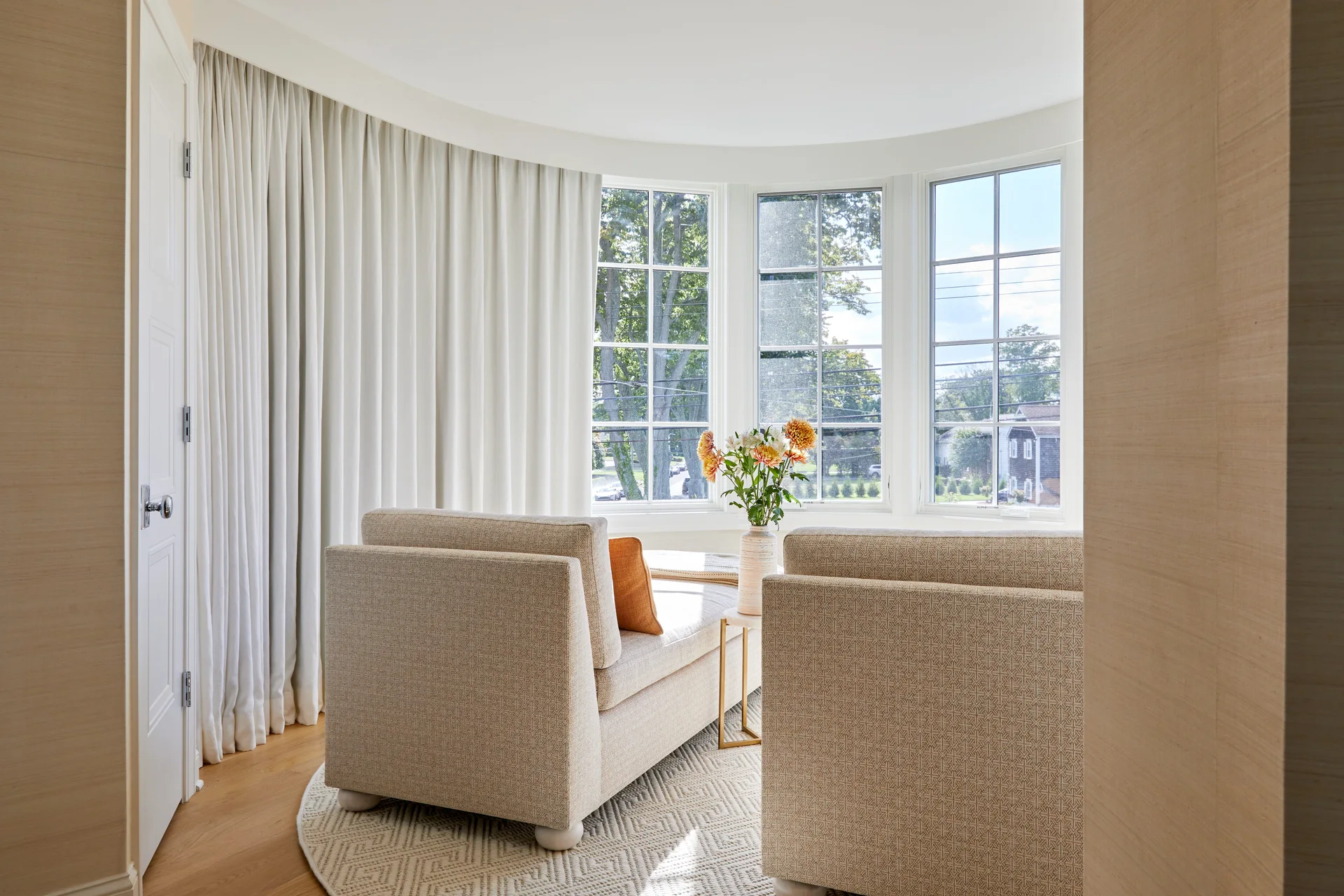 A sunny sitting area featuring two patterned beige lounge chairs placed on a geometric rug in front of a curved bay window with white curtains.