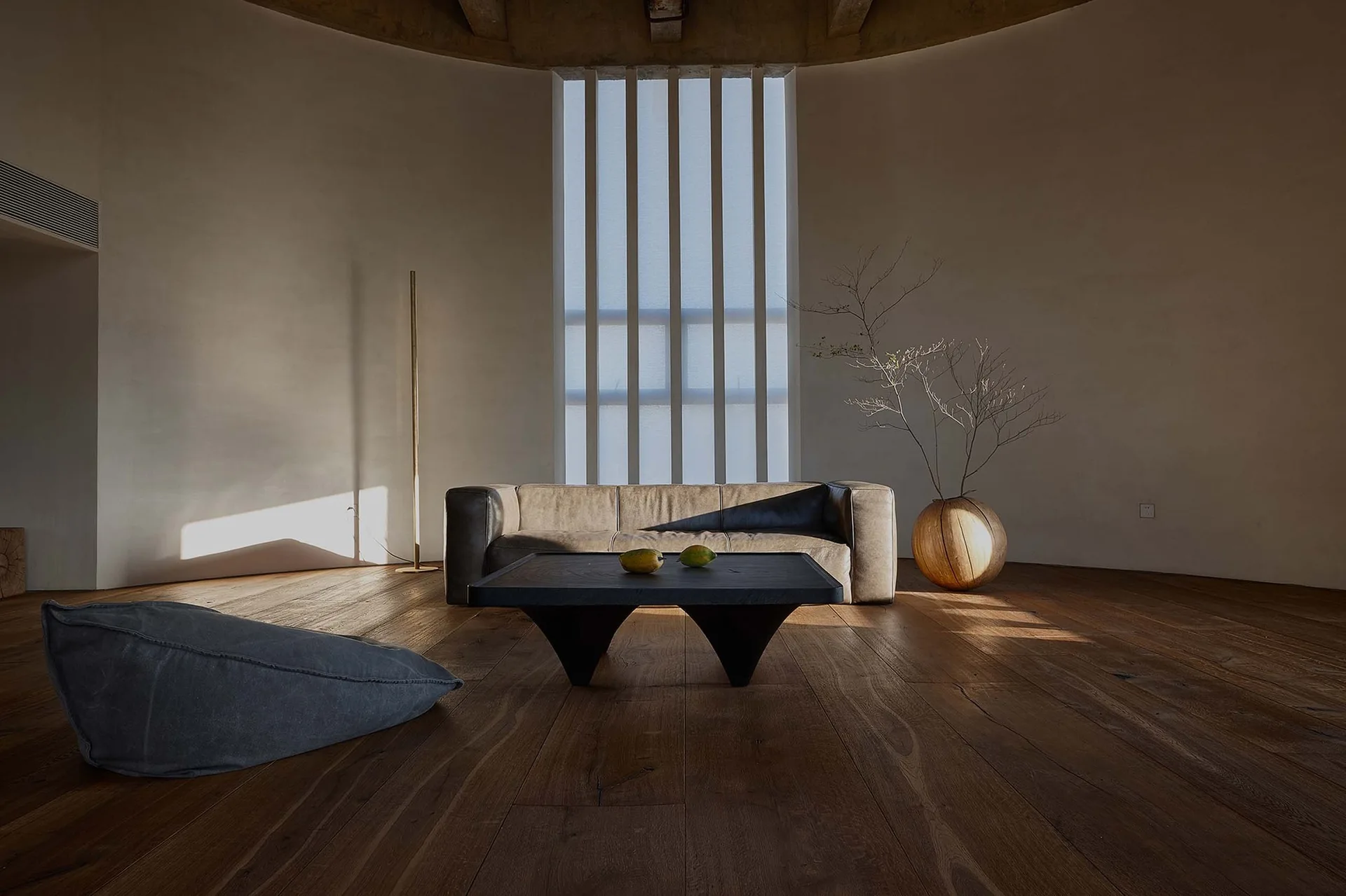 A low grey leather sofa and black wooden coffee table sit on wide-plank floors inside a minimalist curved room with plaster walls.