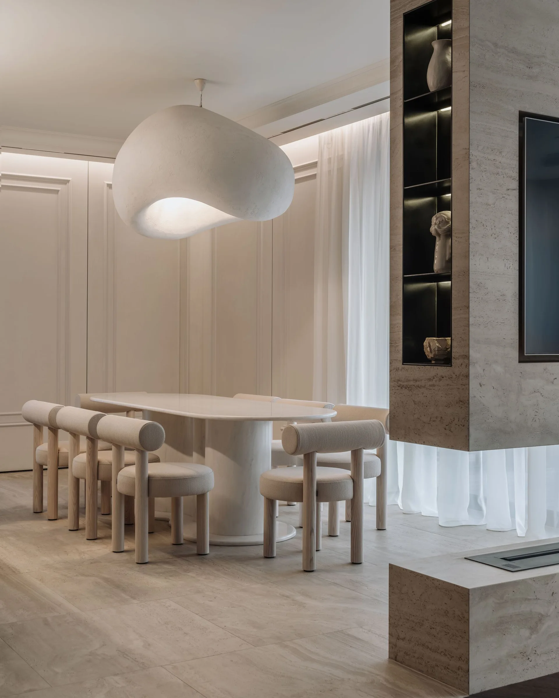 A minimalist cream dining room features an oval table, cylinder-legged chairs, a large biomorphic pendant light, and a stone fireplace with built-in shelves.