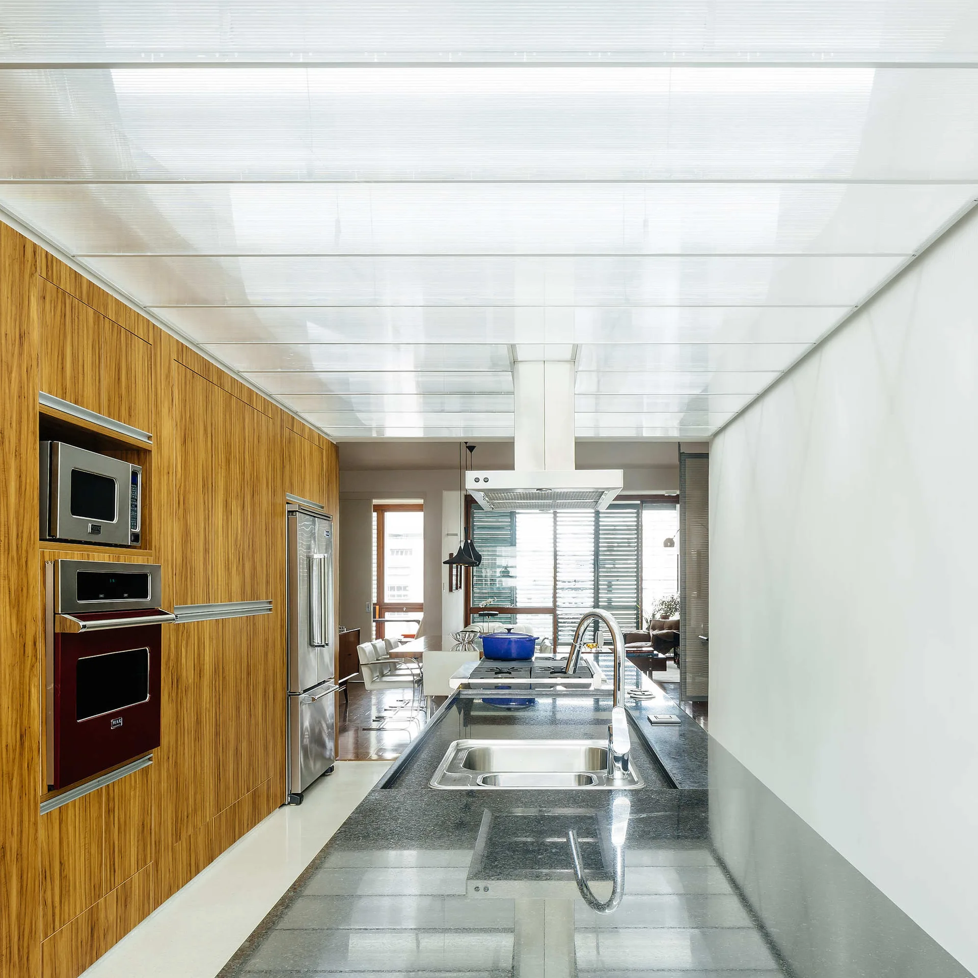 A modern kitchen featuring a long grey stone island with a sink, a full-height wood cabinetry wall, and a large backlit translucent ceiling panel.