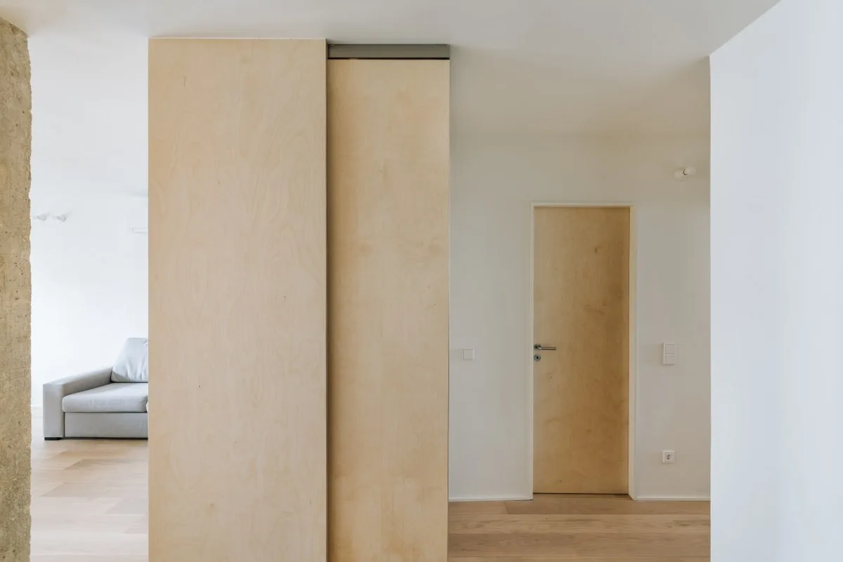 A minimalist hallway features a tall plywood sliding door, a matching wooden room door, white walls, and light wood plank flooring.