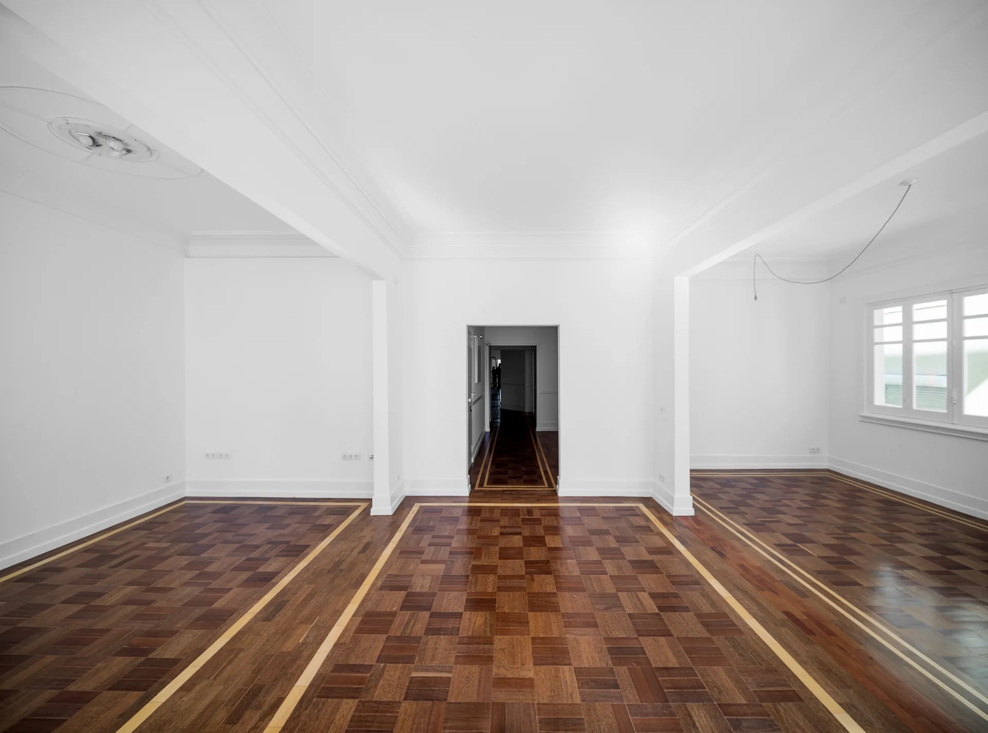 An empty white room features dark brown parquet wood floors with light wood inlay borders, crown moldings, and an open doorway leading to a hallway.