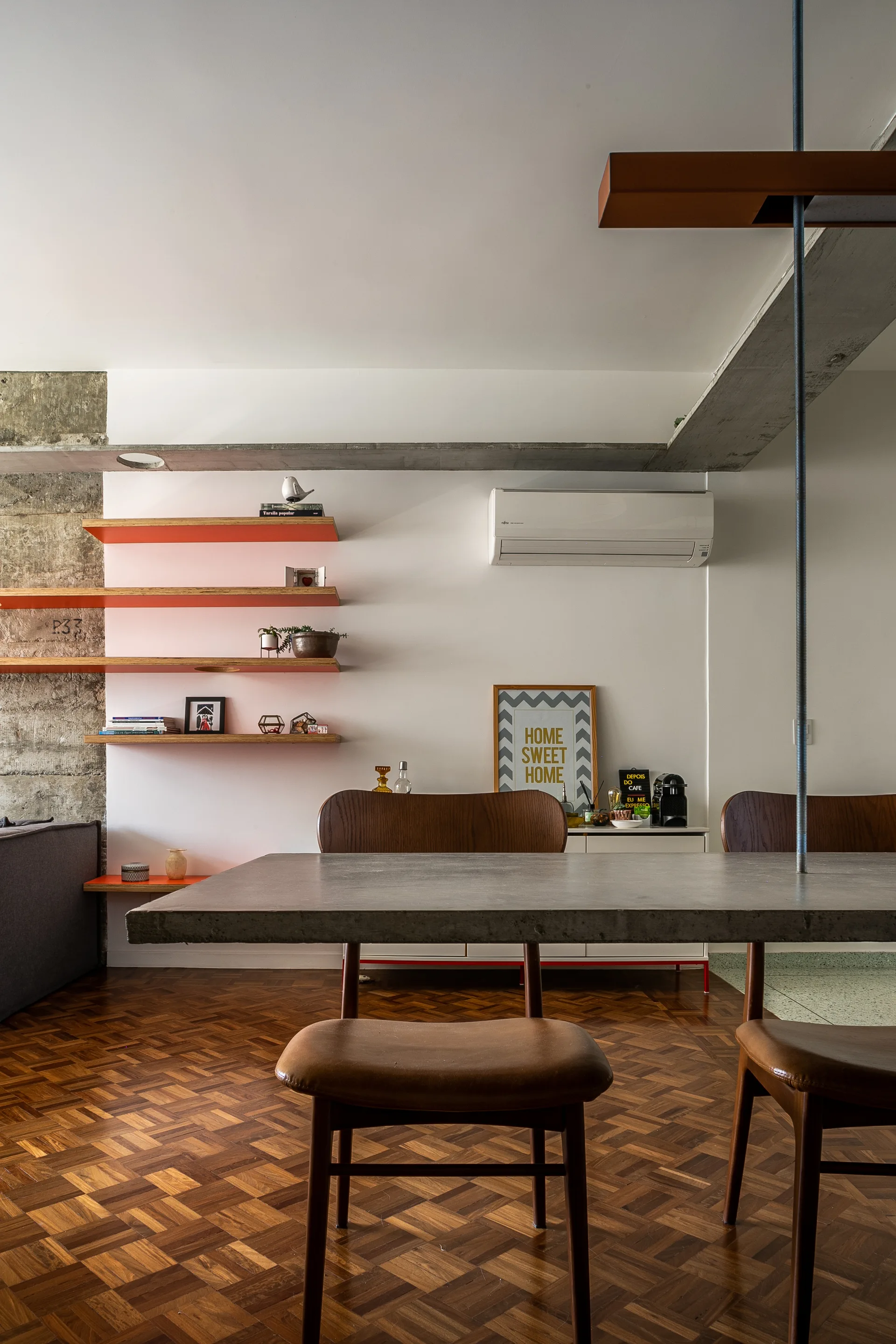 A modern dining room featuring a concrete-topped table, wood chairs, parquet flooring, and orange floating shelves on a white wall.