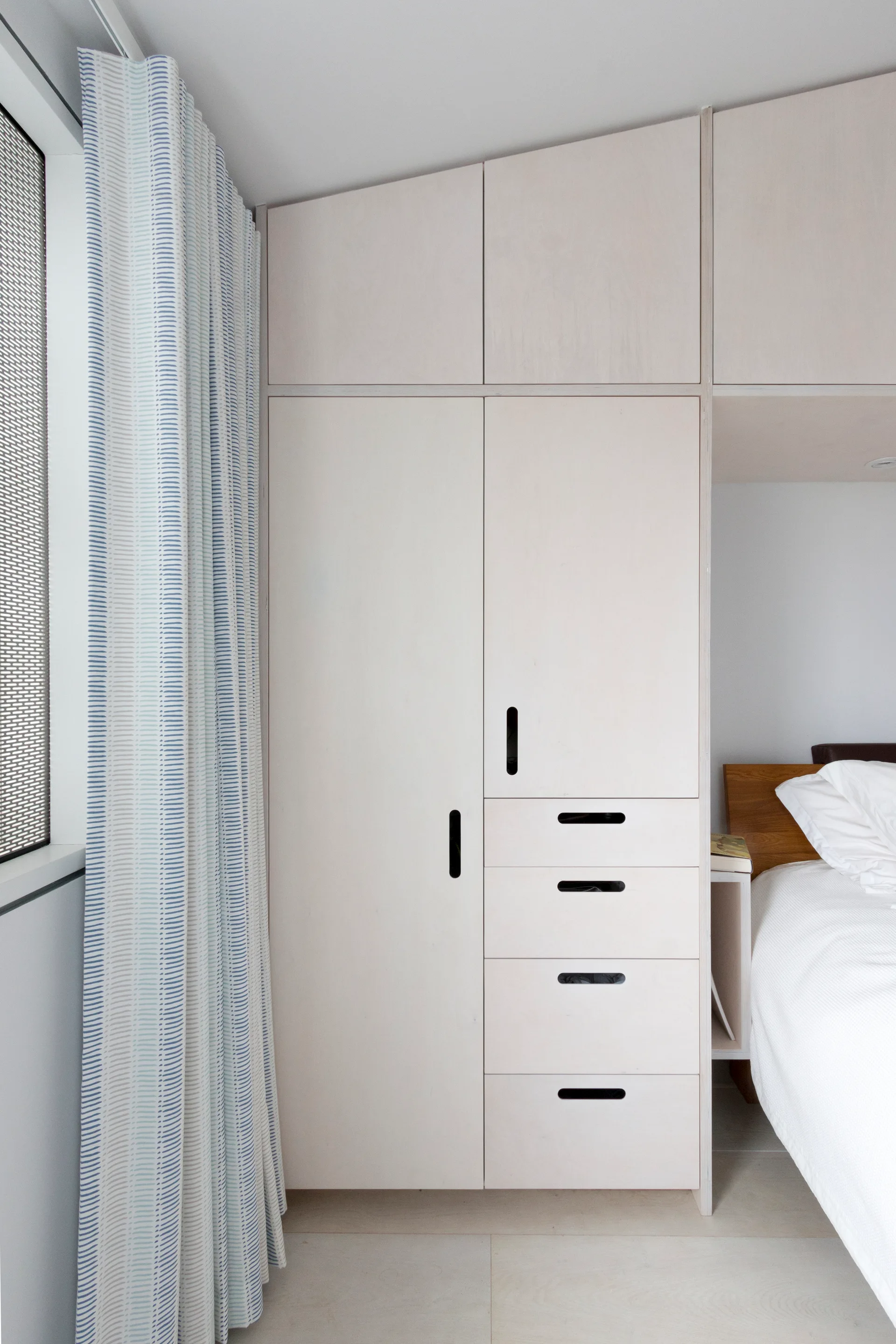 A light wood built-in wardrobe with recessed cut-out handles stands next to a bed and a blue striped floor-to-ceiling curtain.