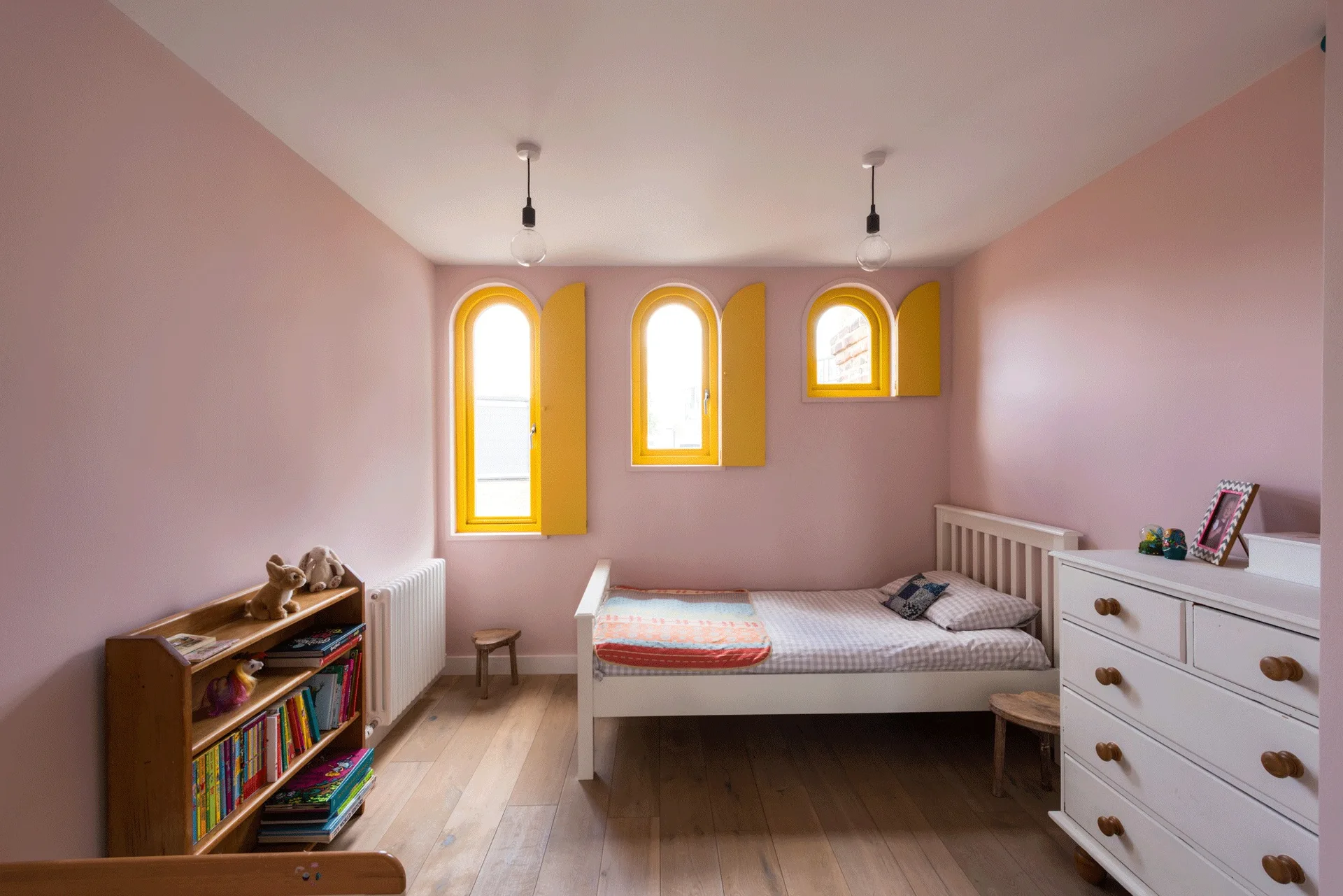 A child's bedroom with pink walls, light wood flooring, a white single bed, and three arched windows fitted with bright yellow shutters.