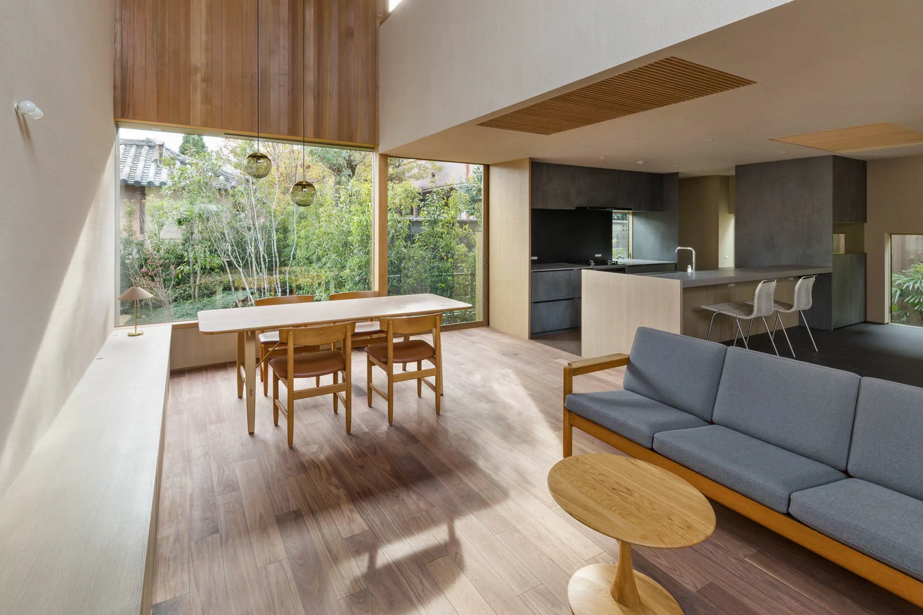 A modern open-plan living and dining room with a grey sofa, wooden table, and a tall window showing garden greenery.