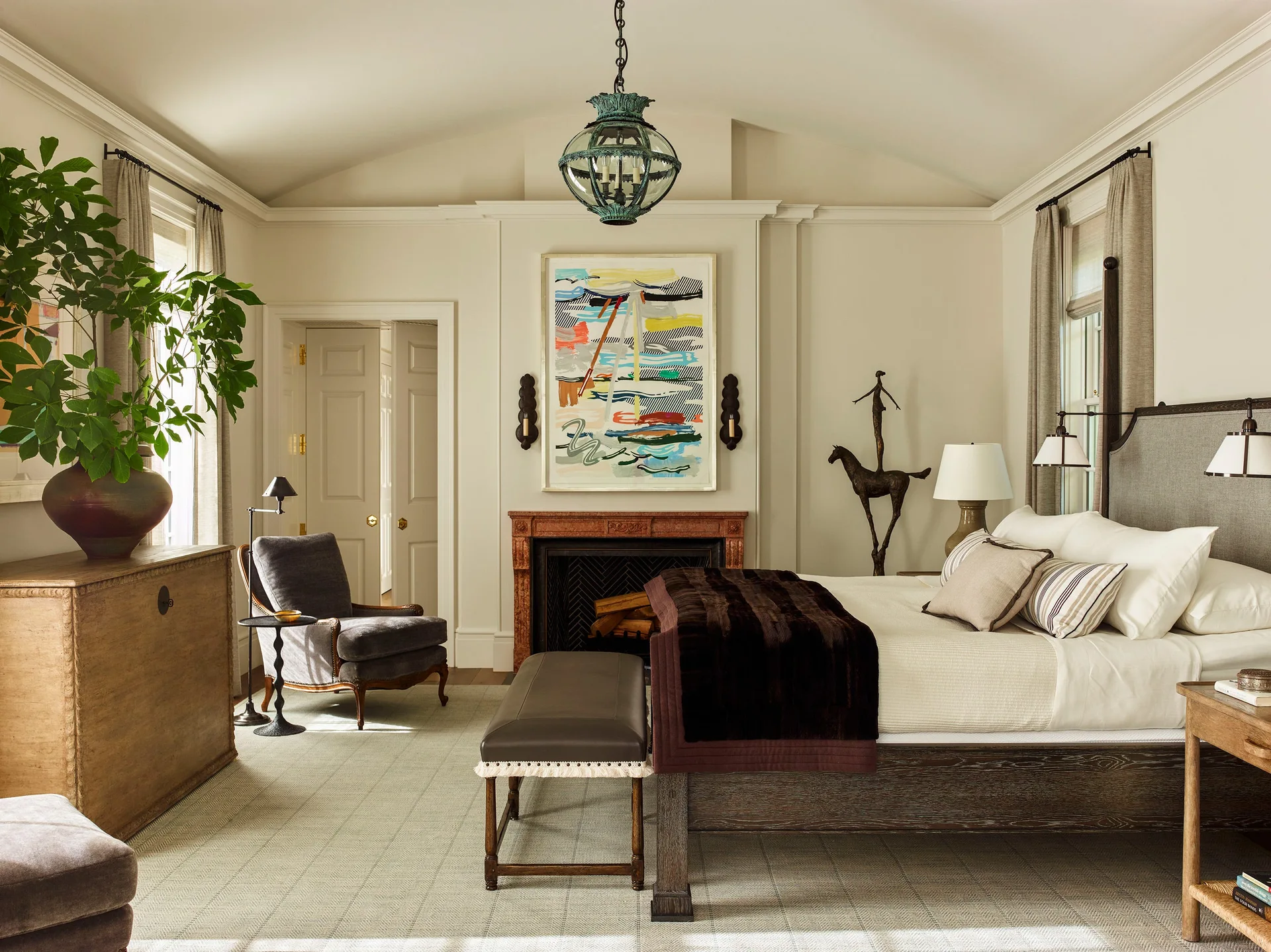 A bedroom features a vaulted ceiling, a stone fireplace with abstract art above it, and an upholstered bed with a dark throw.