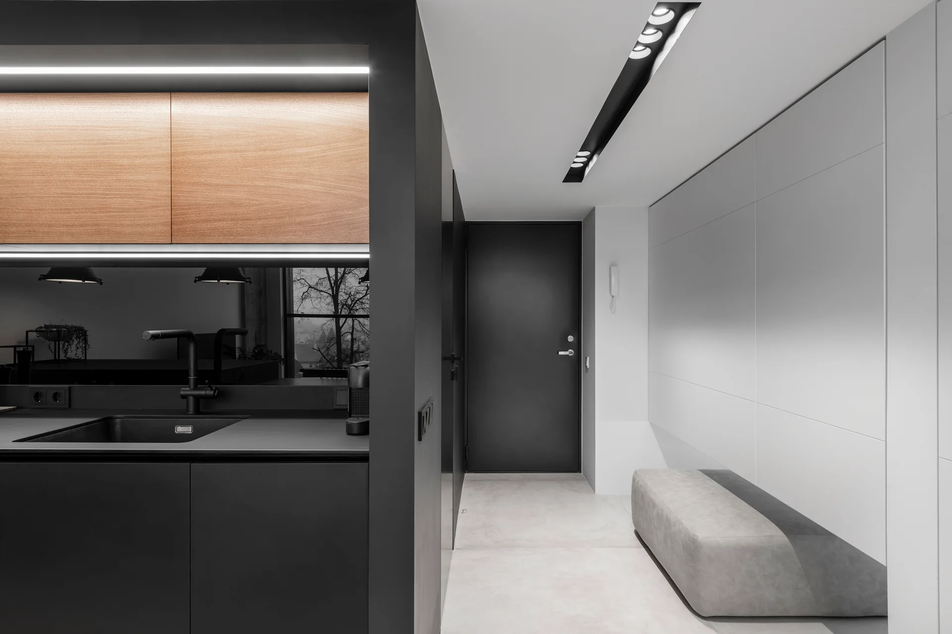 A minimalist black kitchen counter with an integrated sink and wood upper cabinet sits adjacent to a bright hallway featuring a grey bench.