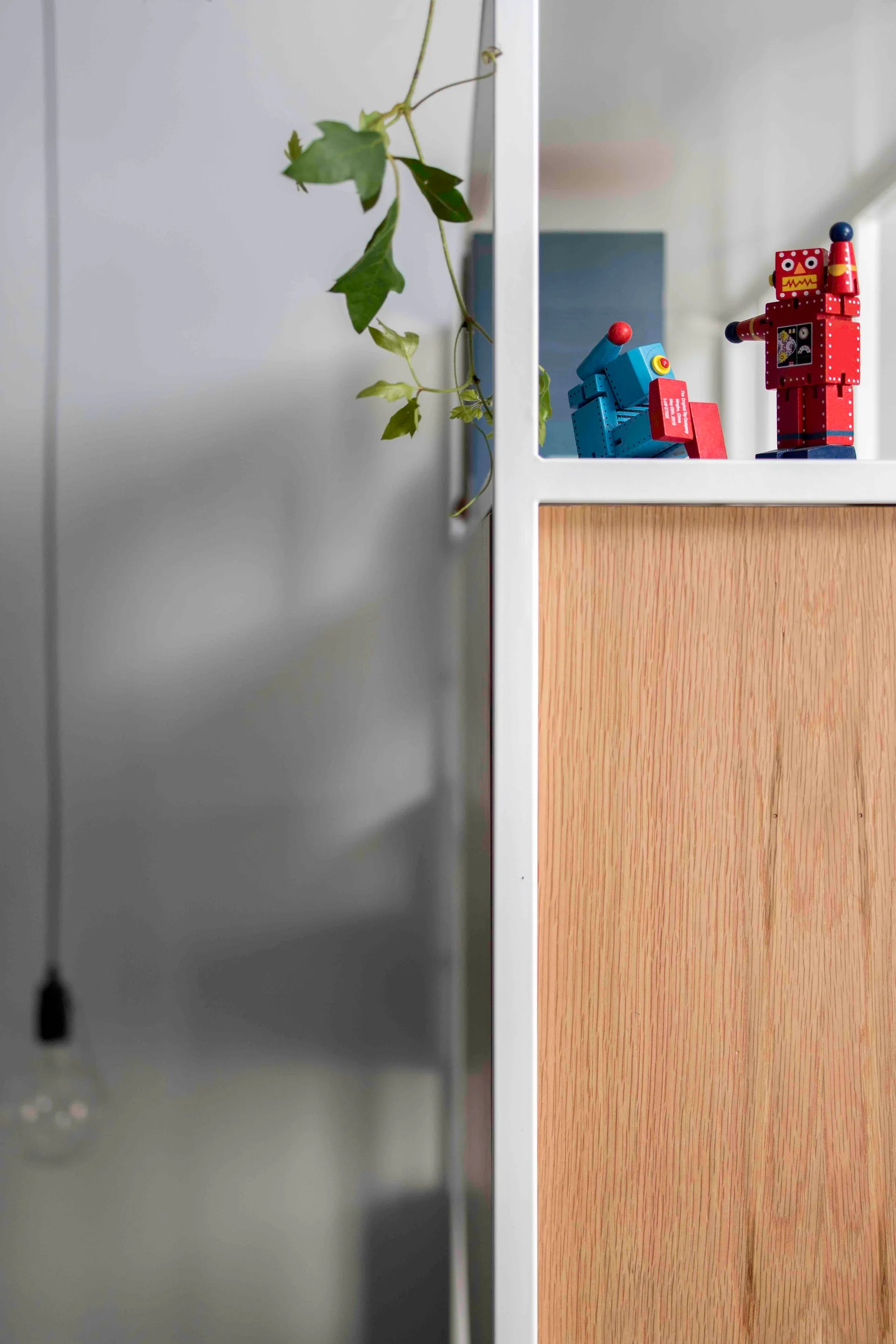 A close-up of a white metal and wood shelving unit decorated with two small robot toys and a trailing green vine against a gray background.