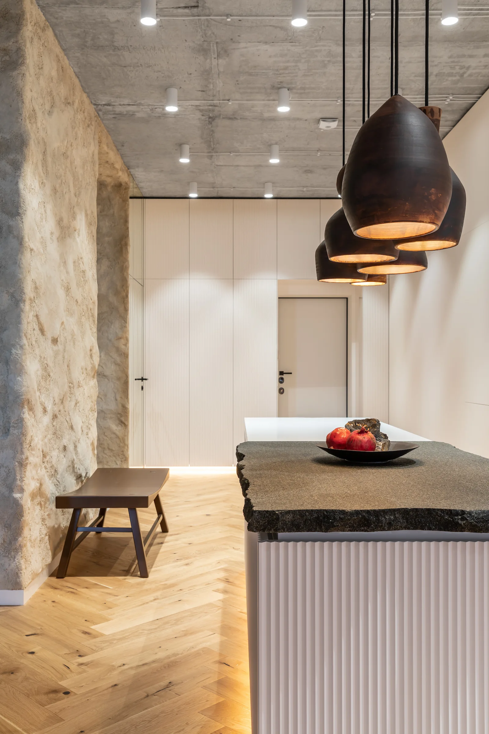 A modern kitchen featuring a textured stone accent wall, a white fluted island with a dark stone countertop, and herringbone wood floors.