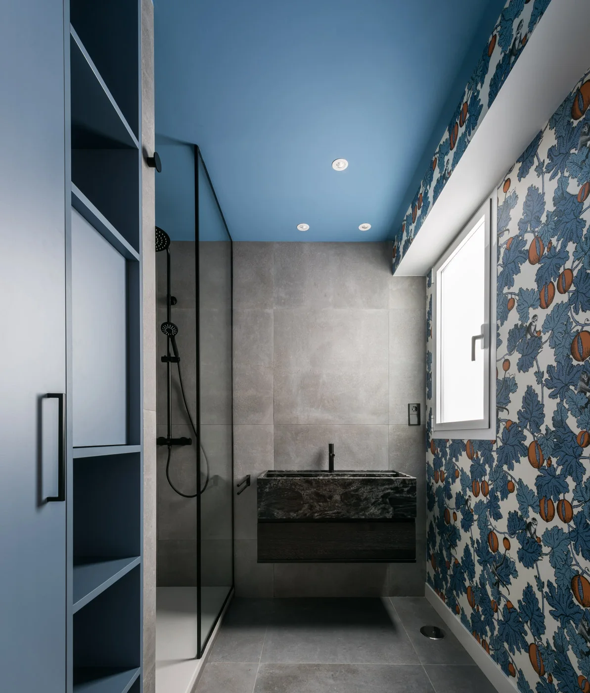 A modern bathroom features a bold blue ceiling, fruit-patterned wallpaper, a floating dark stone vanity, and grey tiled walls.