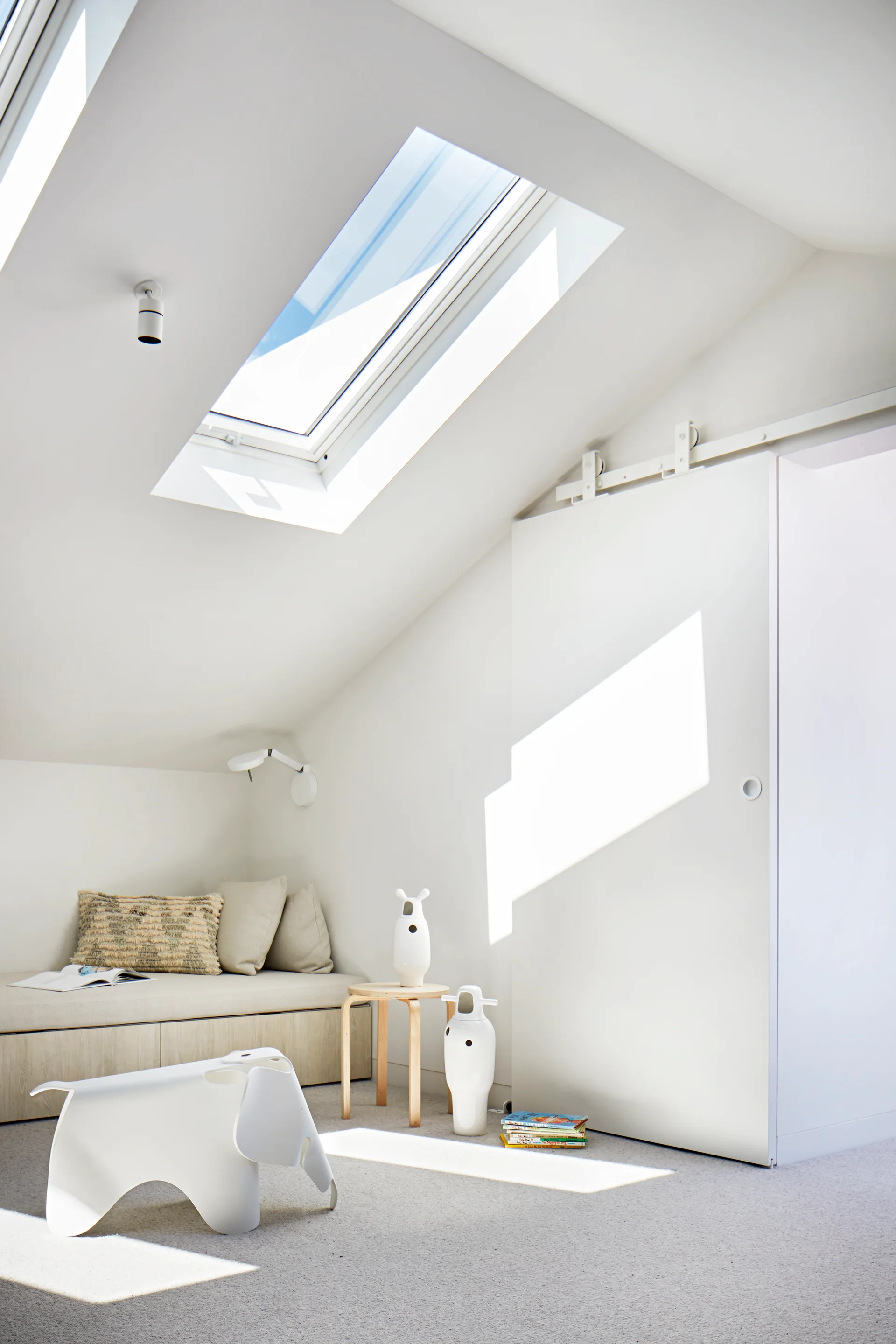 A minimalist attic bedroom with a sloped ceiling and skylight features a built-in daybed, a white sliding door, and a wooden side table.