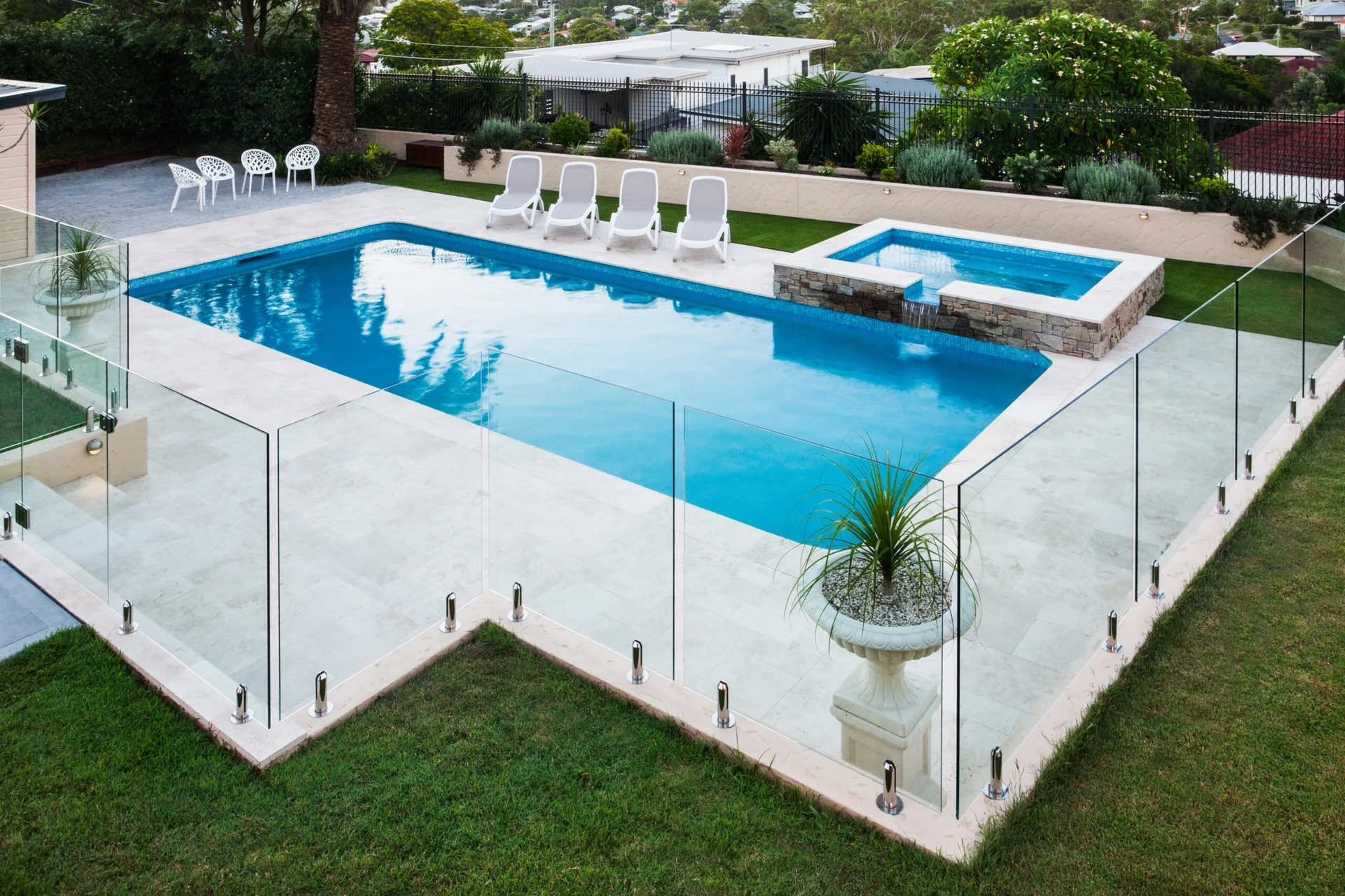 A rectangular swimming pool and integrated spa are surrounded by light stone paving and a frameless glass safety fence in a backyard.