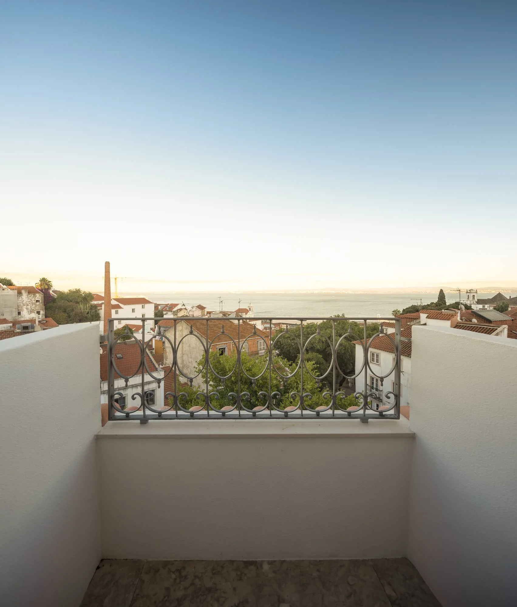 A small balcony with plain white stucco walls, mottled stone floor tiles, and a decorative wrought iron railing overlooking terracotta rooftops.