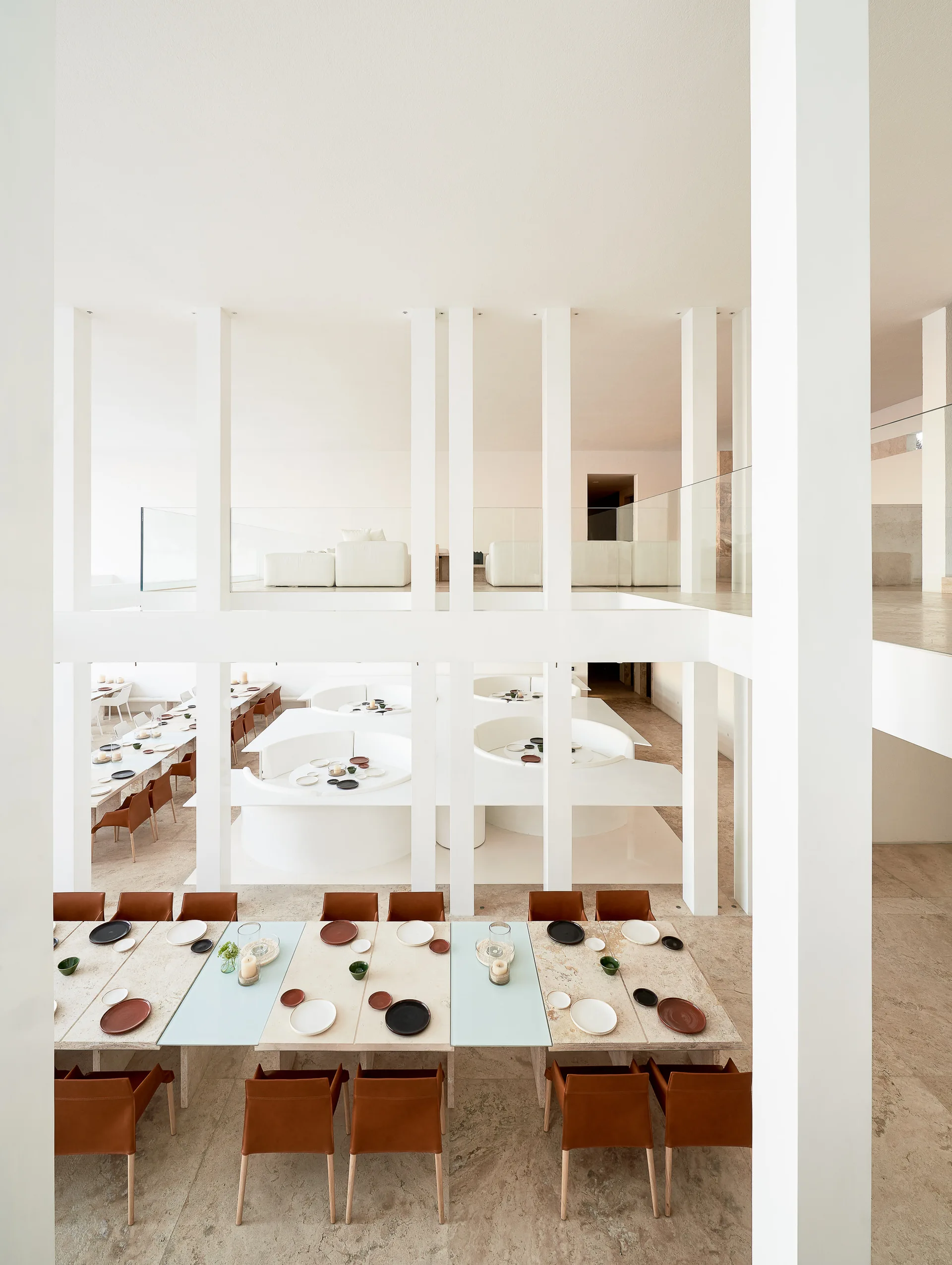 A multi-level dining hall features long stone tables with tan leather chairs arranged between prominent white structural columns.