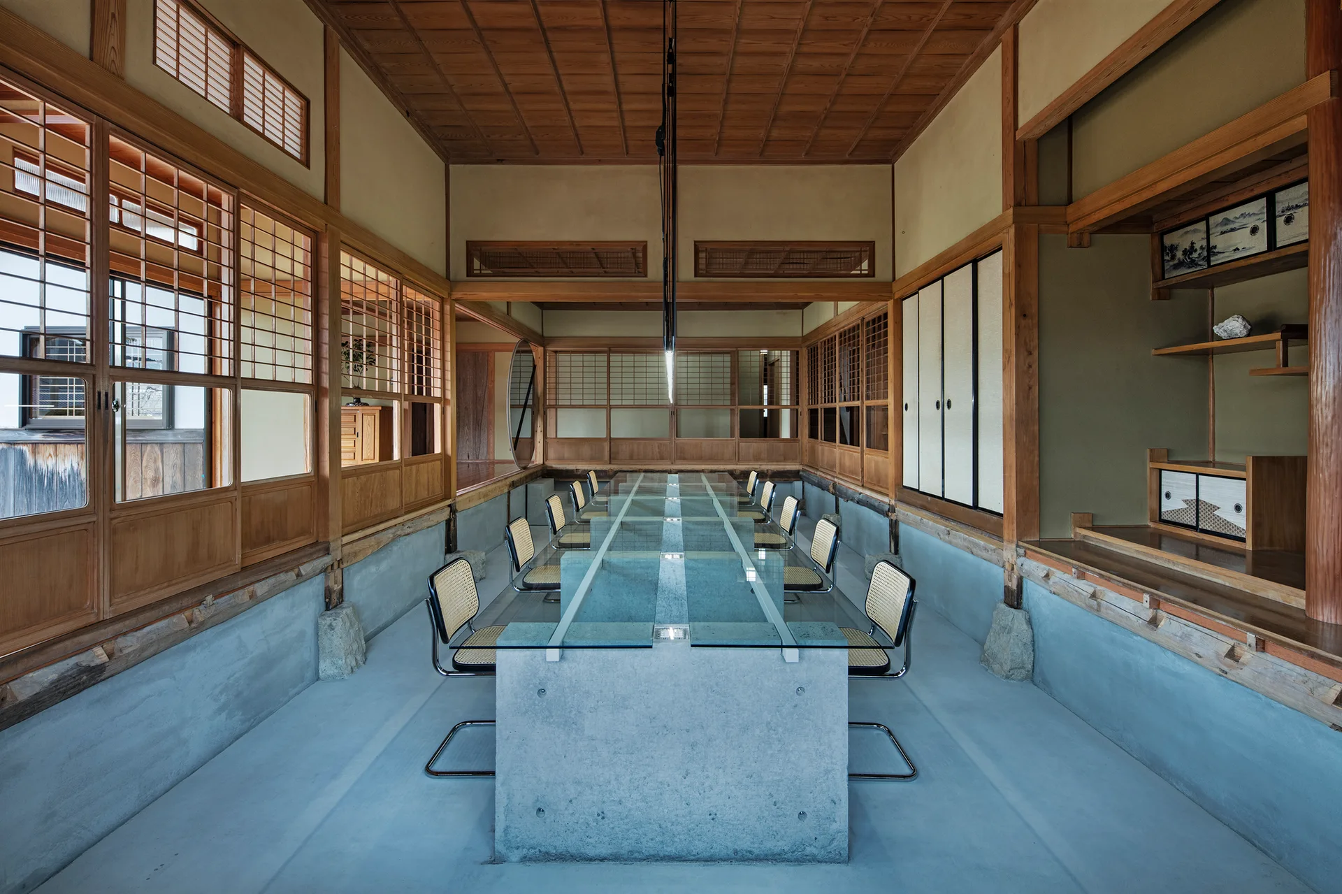 A long glass table with a concrete base and cane chairs sits inside a traditional Japanese room with wood ceilings and shoji screens.