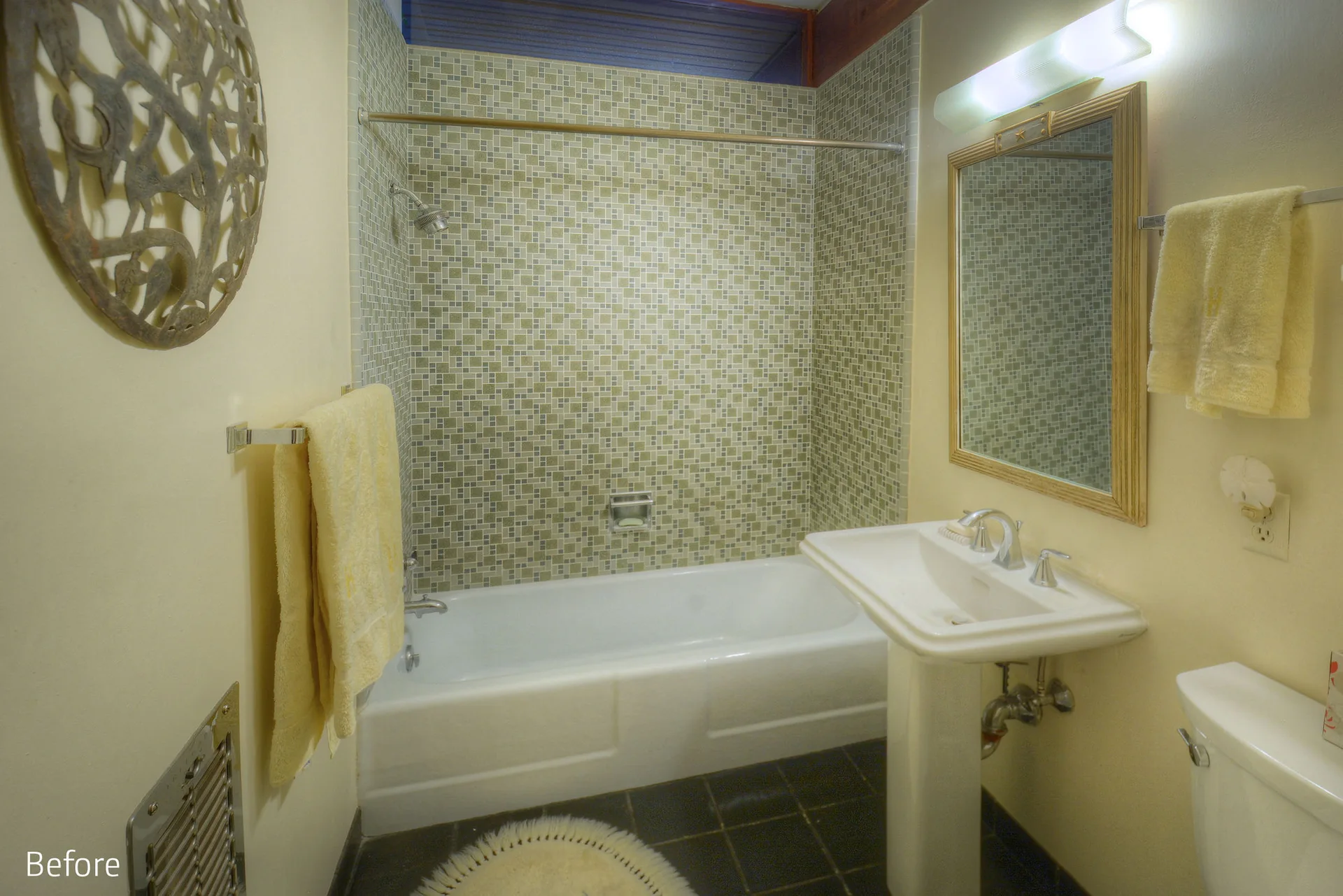A small bathroom features a white pedestal sink, a bathtub with green mosaic wall tiles, and yellow towels hanging on chrome bars.