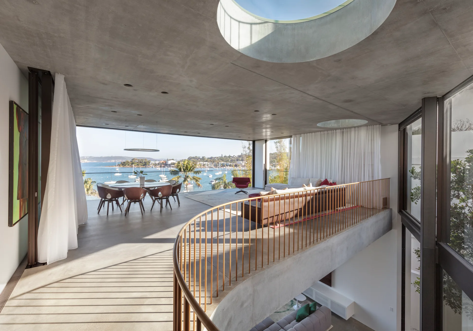 A concrete mezzanine living and dining space featuring a curved metal railing, circular skylights, and expansive harbor views.