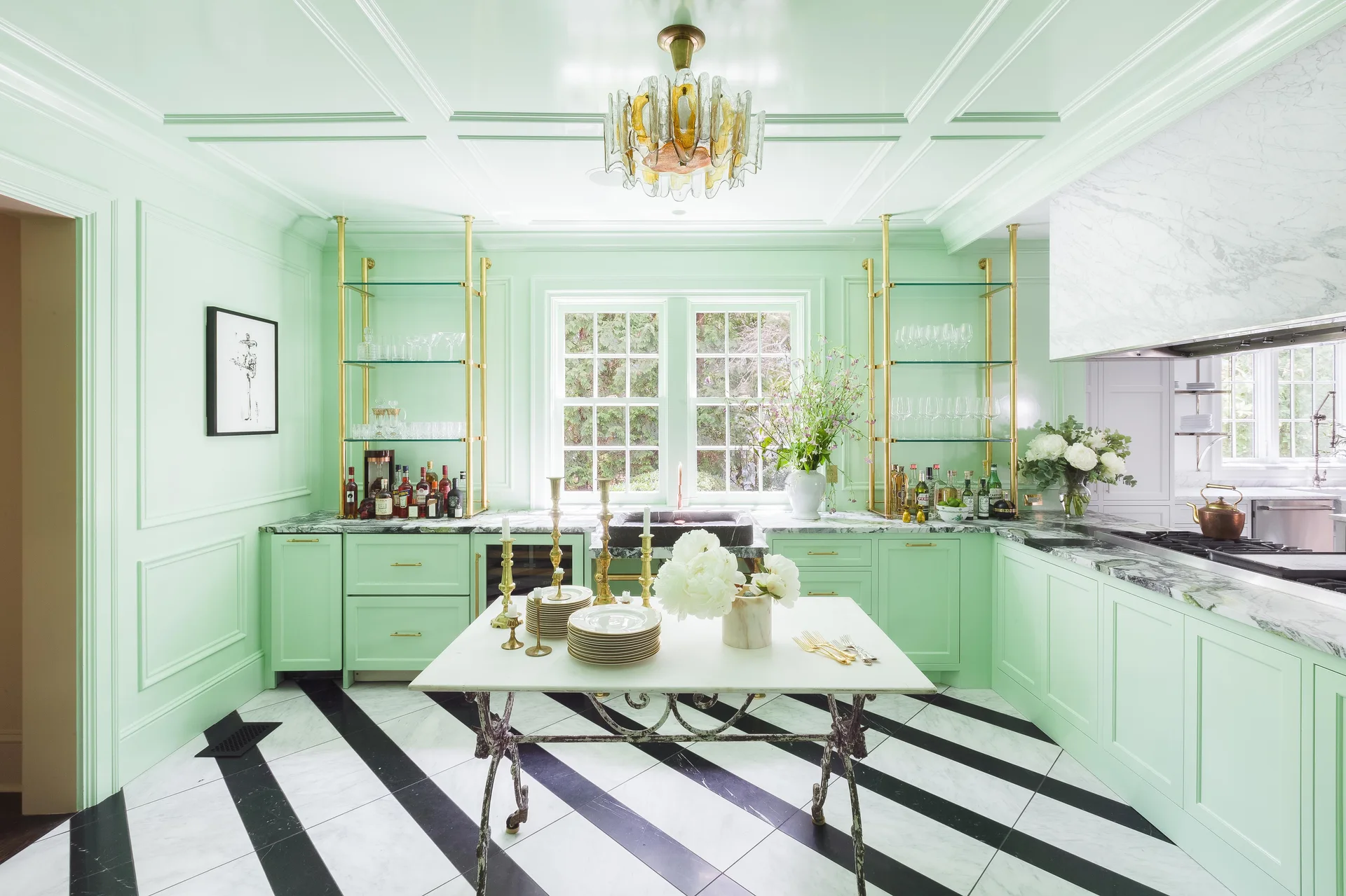 A mint green kitchen features glossy paneled walls, diagonal black and white marble floor tiles, brass shelving units, and a central table island.