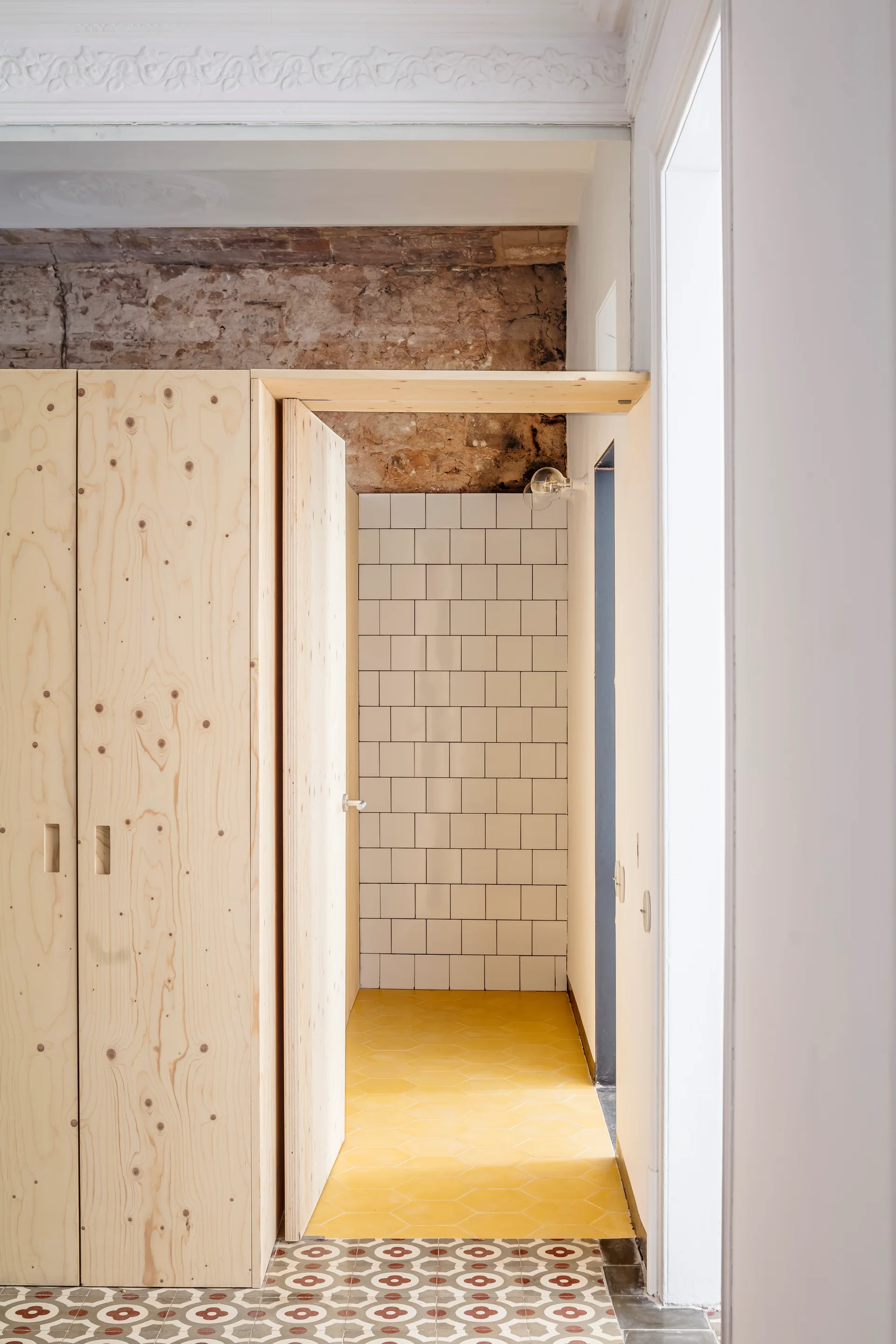 A transitional space features light plywood cabinetry, a white tiled wall under exposed brick, and yellow hexagonal floor tiles.