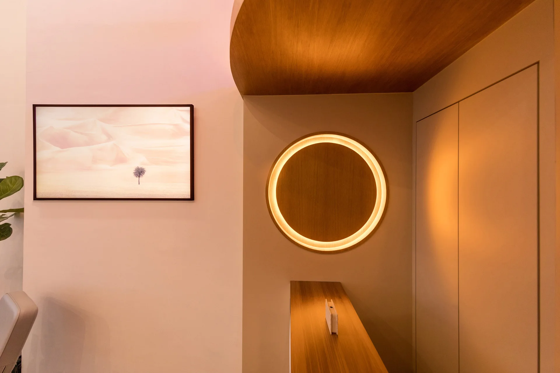 A minimalist beige wall displays a circular backlit wood panel next to framed desert art, with a curved wooden ceiling and built-in cabinets.