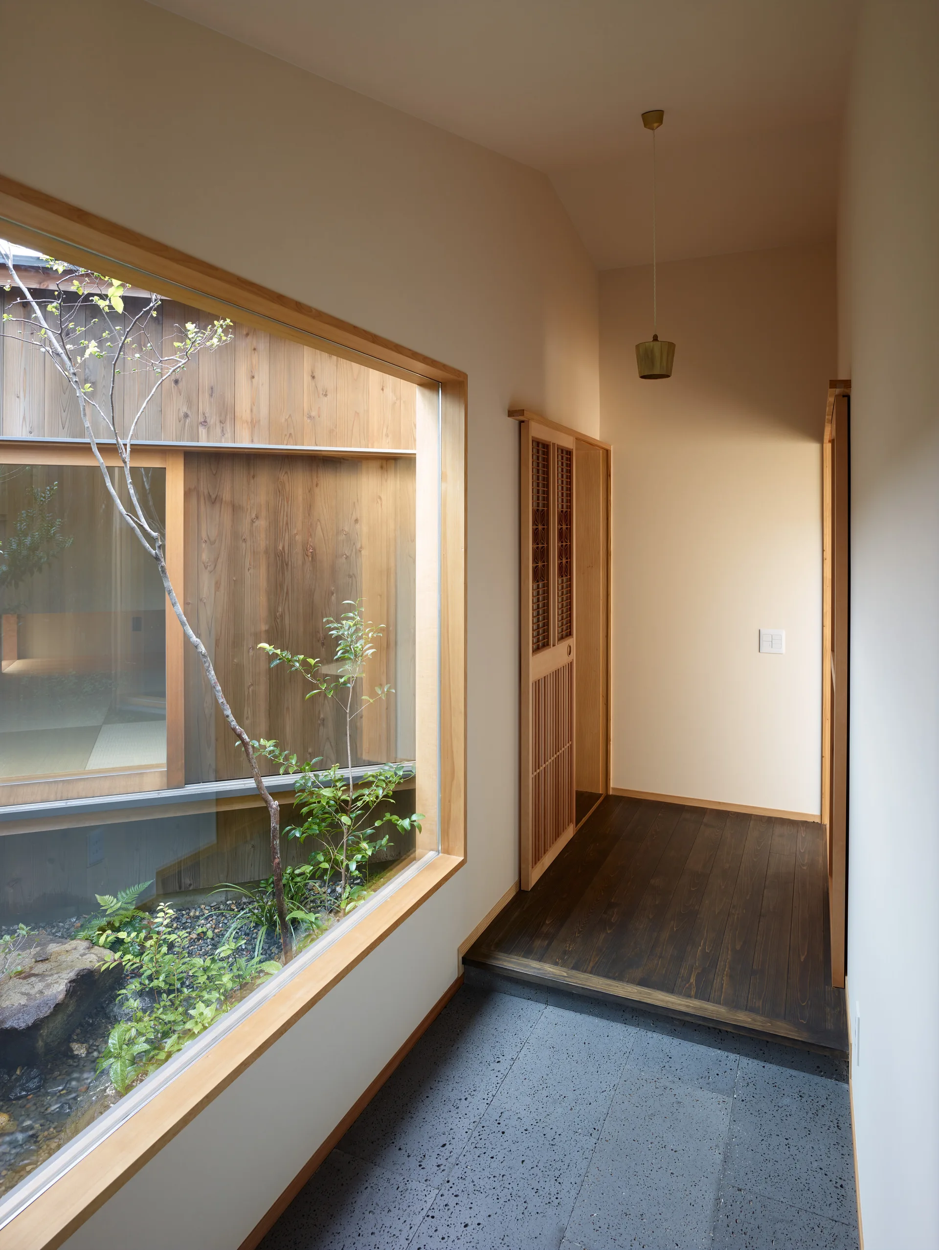 A minimalist hallway transitions from textured grey stone tiles to dark wood flooring beside a large window looking out onto a small garden.