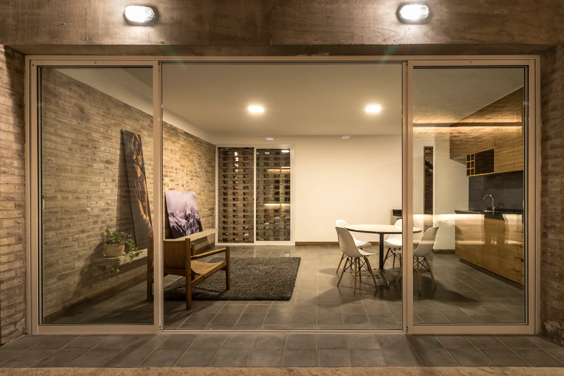 An open-concept living and dining area with an exposed brick wall, tile flooring, and a kitchen seen through large sliding glass doors.