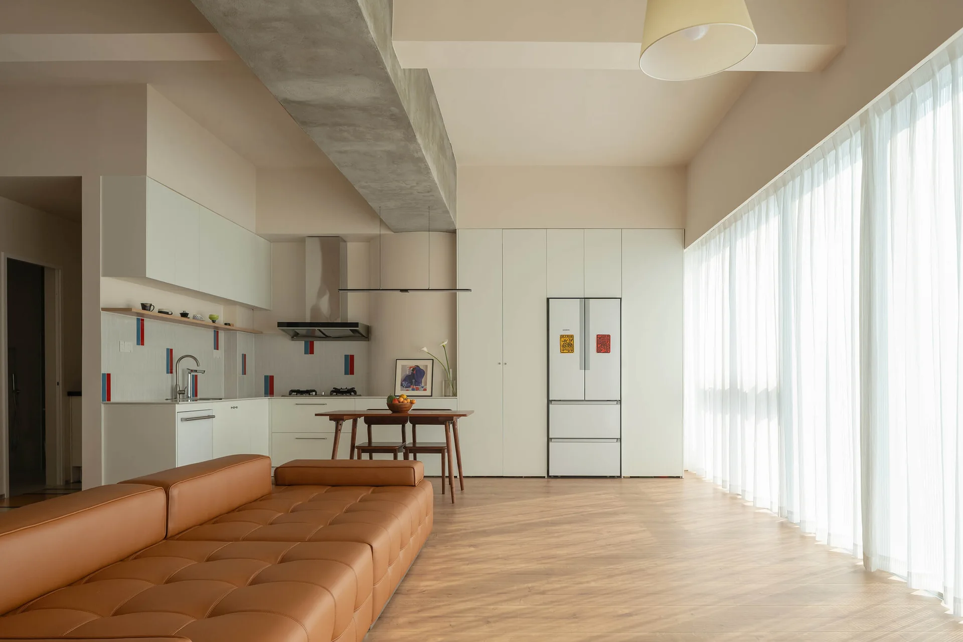 An open-concept living area and kitchen with a tufted tan leather sofa, wood floors, and an exposed concrete ceiling beam.