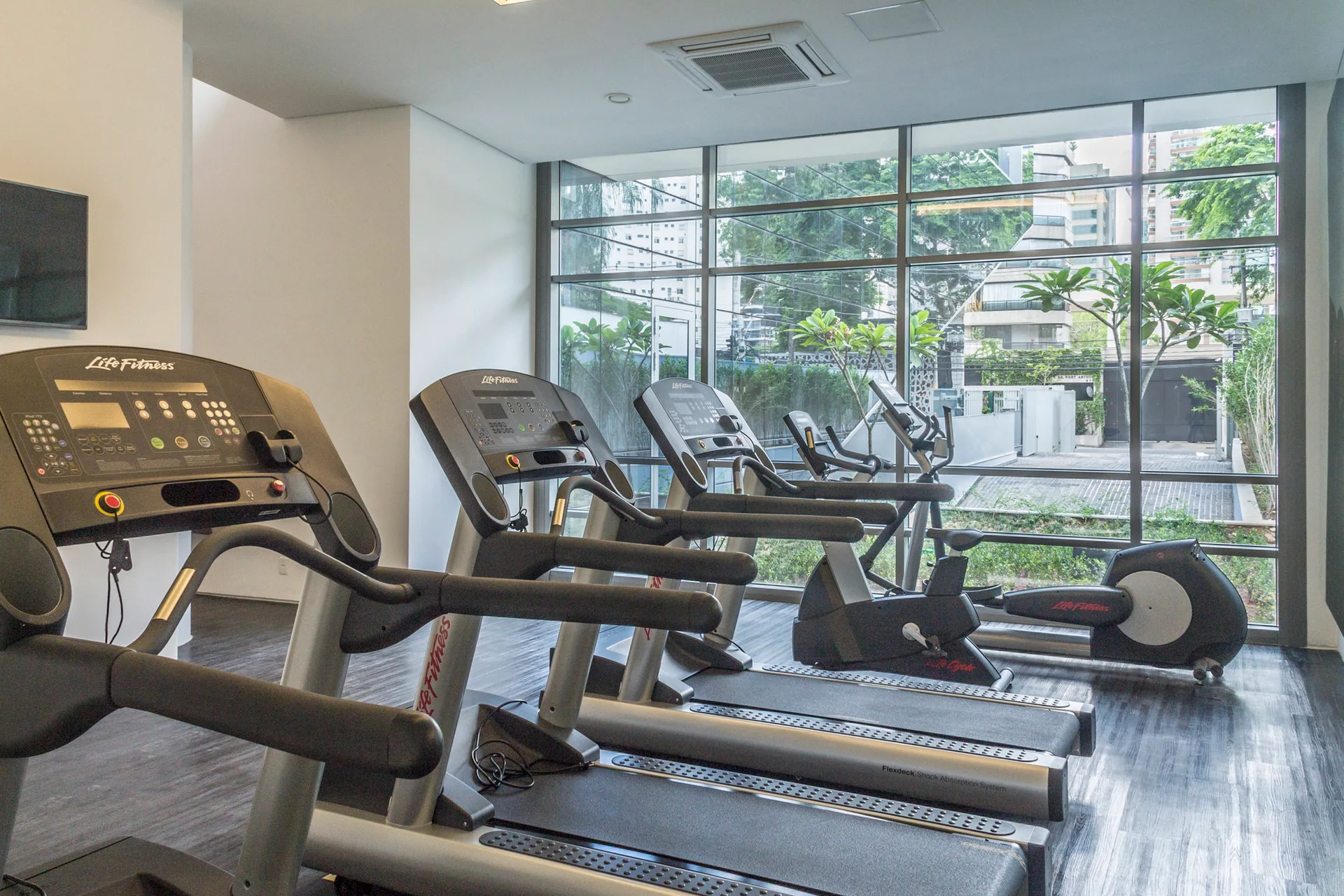 A modern gym interior features a row of treadmills and an exercise bike on dark wood-look flooring next to a large grid window overlooking trees.