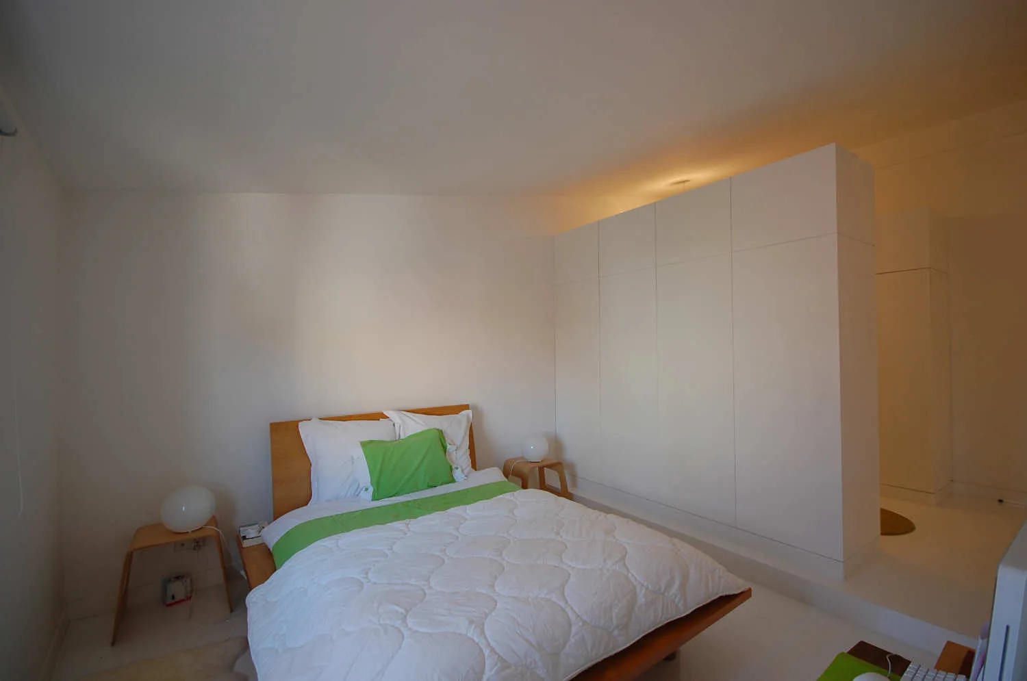 A minimalist bedroom featuring a wooden bed with green pillows next to a large white built-in wardrobe partition wall.
