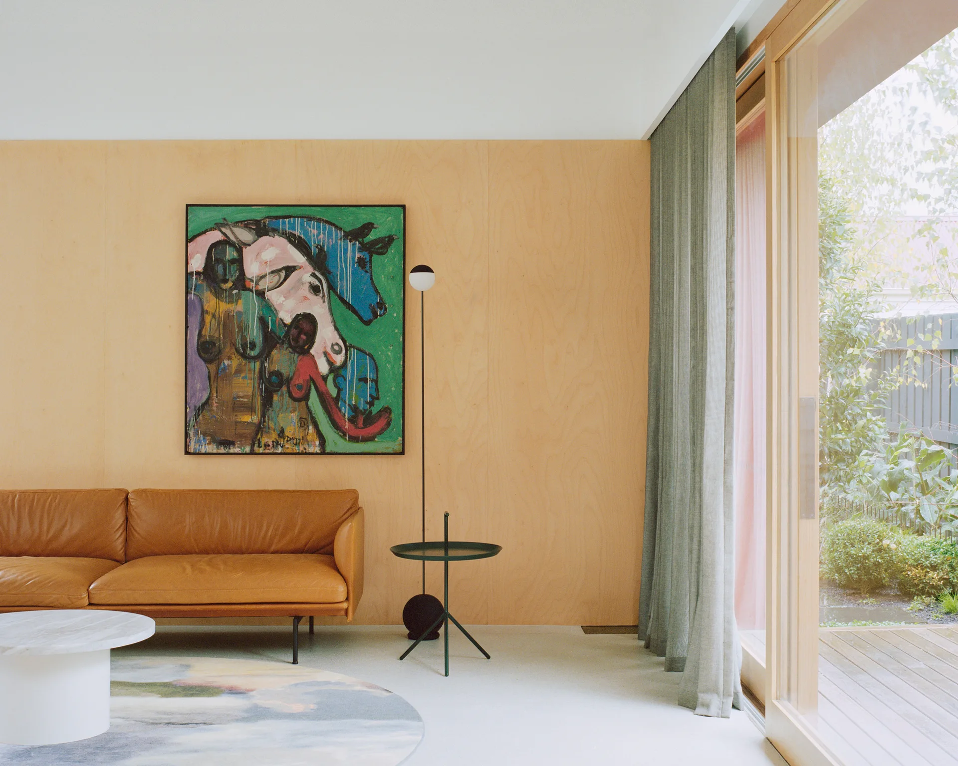 A modern living room features light plywood wall panels, a tan leather sofa, colorful artwork, and a large sliding glass door opening to a garden.