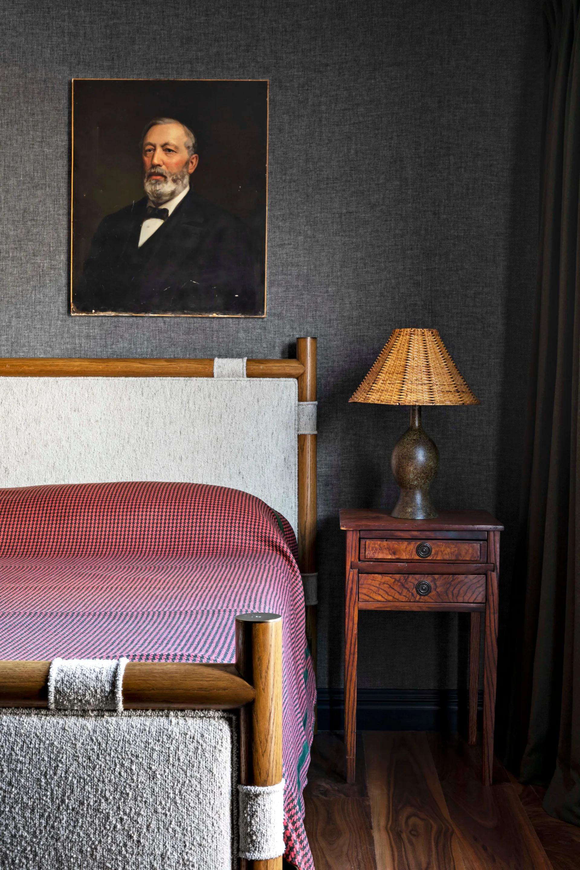 A bedroom corner features a wooden bed with light textured fabric inserts, a dark wood nightstand with a woven lamp, and charcoal wallpaper.