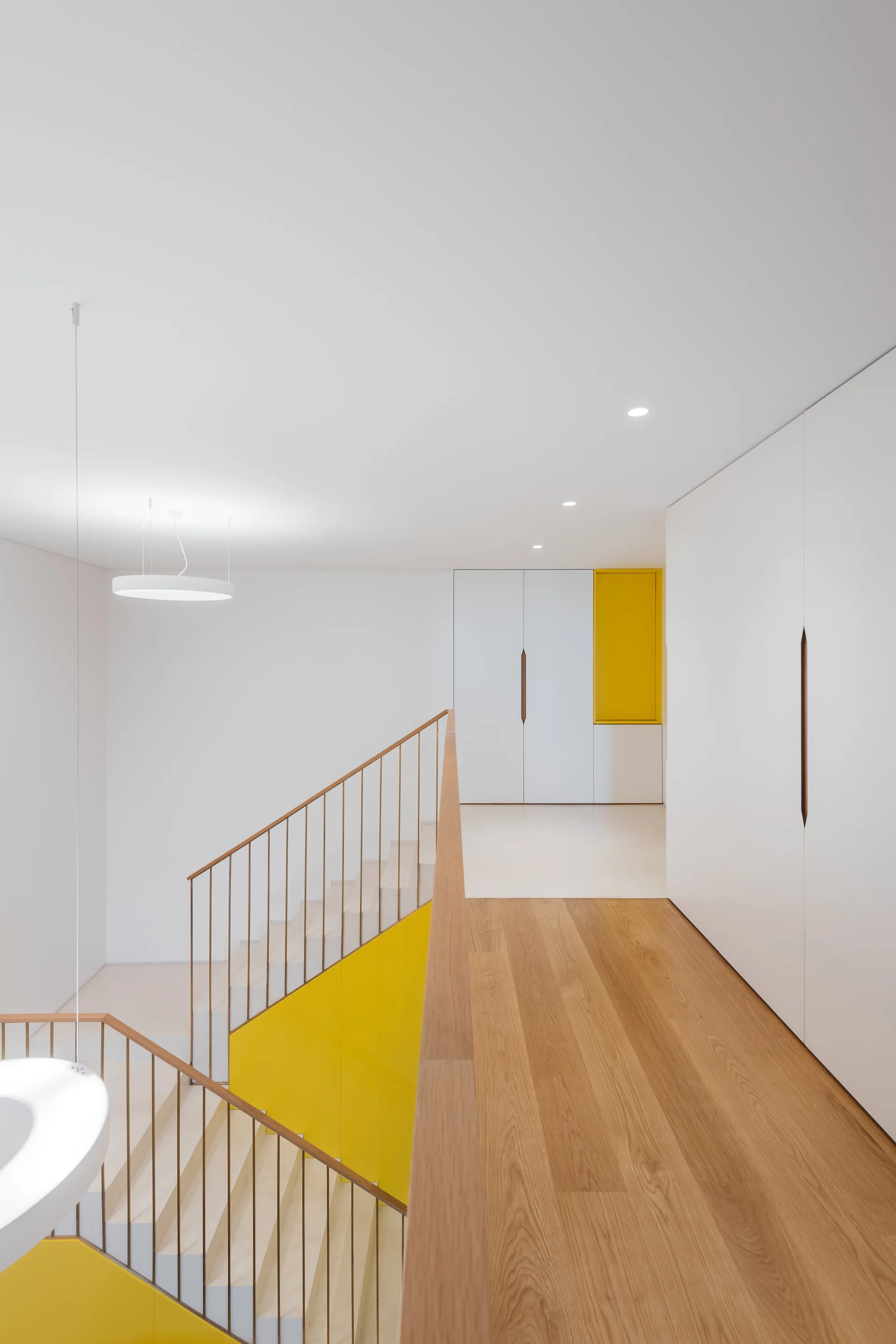 A minimalist hallway and staircase landing with light wood floors, floor-to-ceiling white cabinets, and bright yellow accent panels.